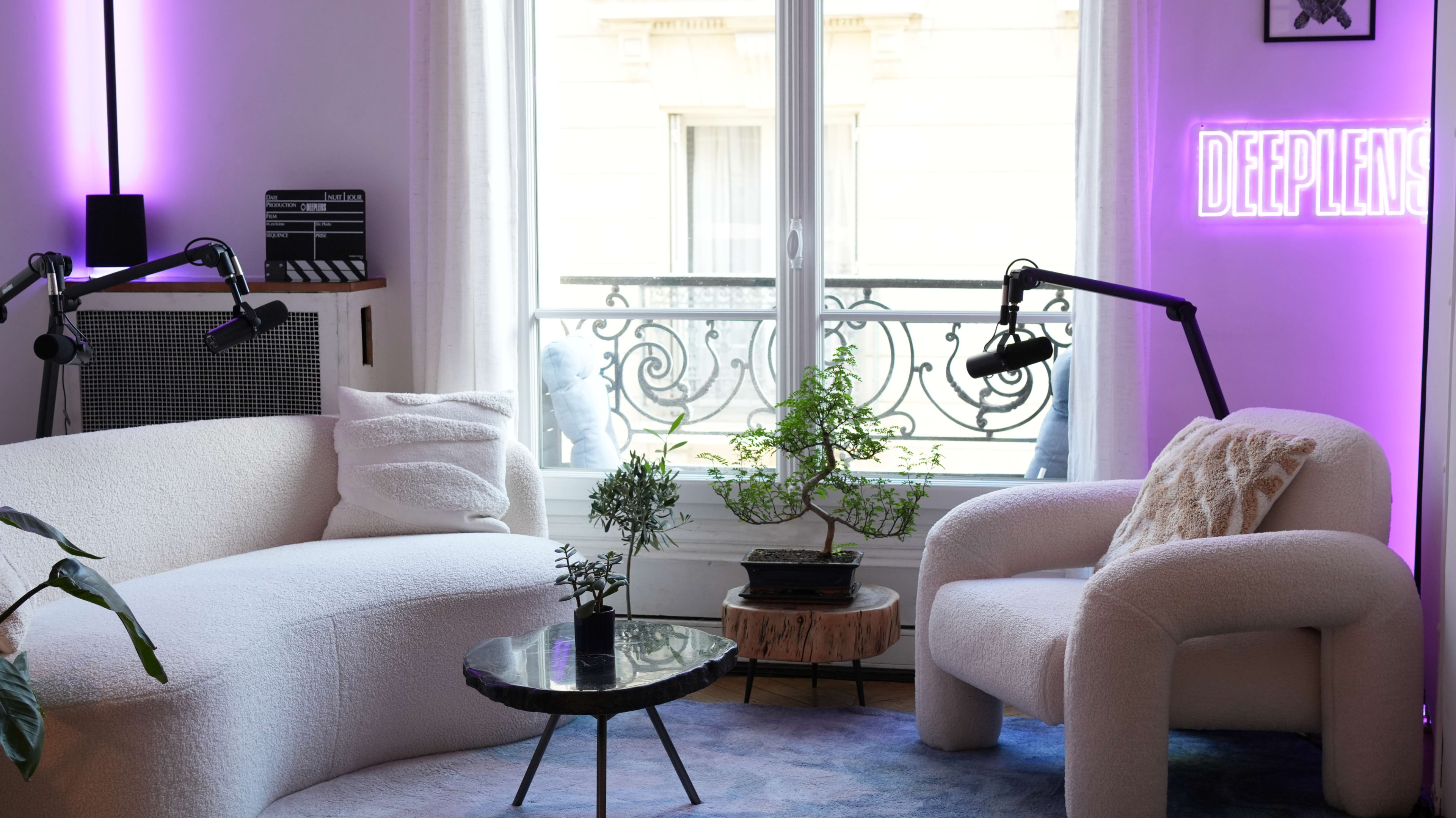 The image shows a modern living room with a white curved sofa and chair, a circular table, and large windows framed by sheer curtains, all illuminated by purple lighting and a neon sign.