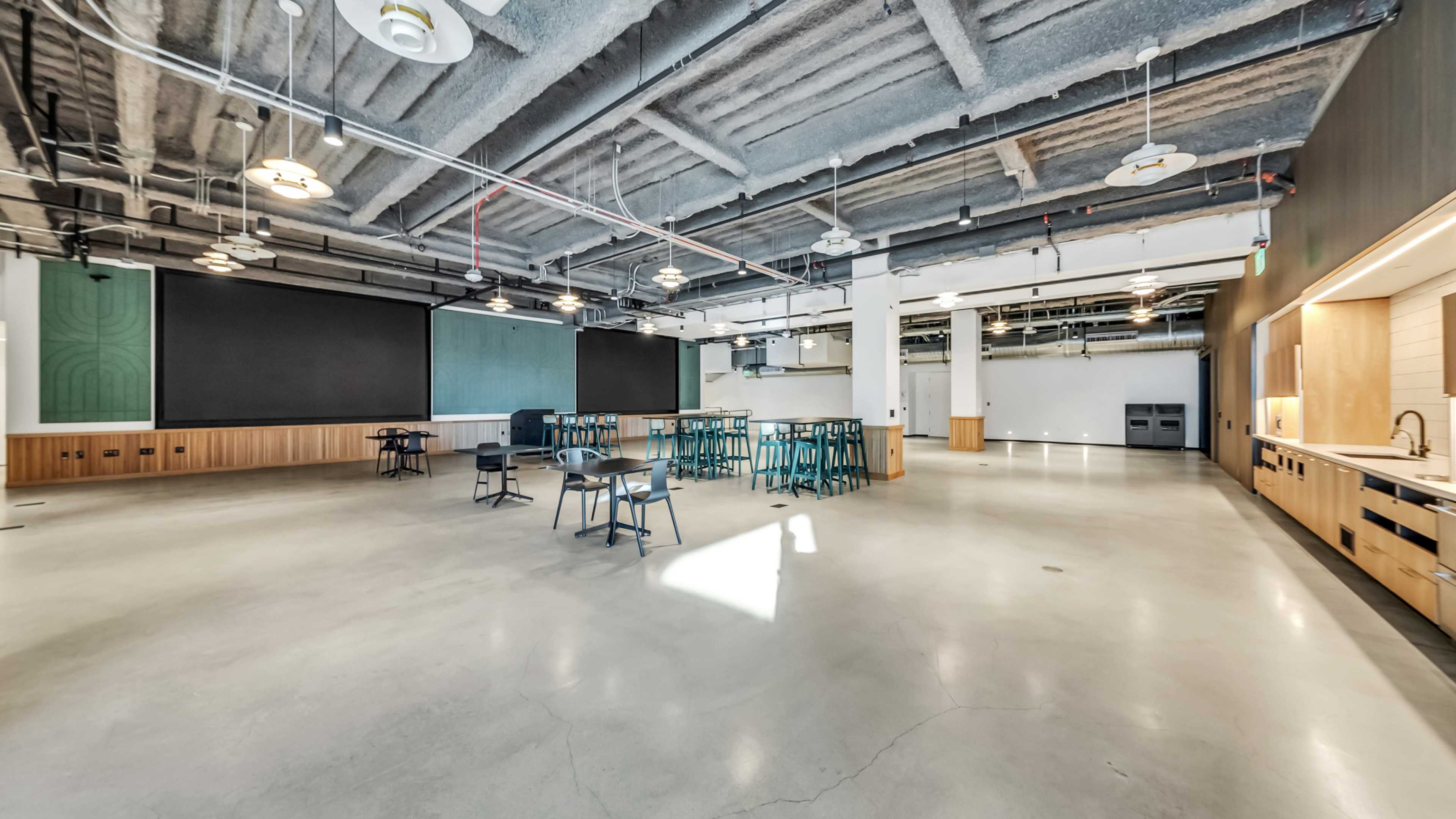 A large, open space with concrete floors, several tables and chairs, and a kitchenette area, all under a high ceiling with visible beams.