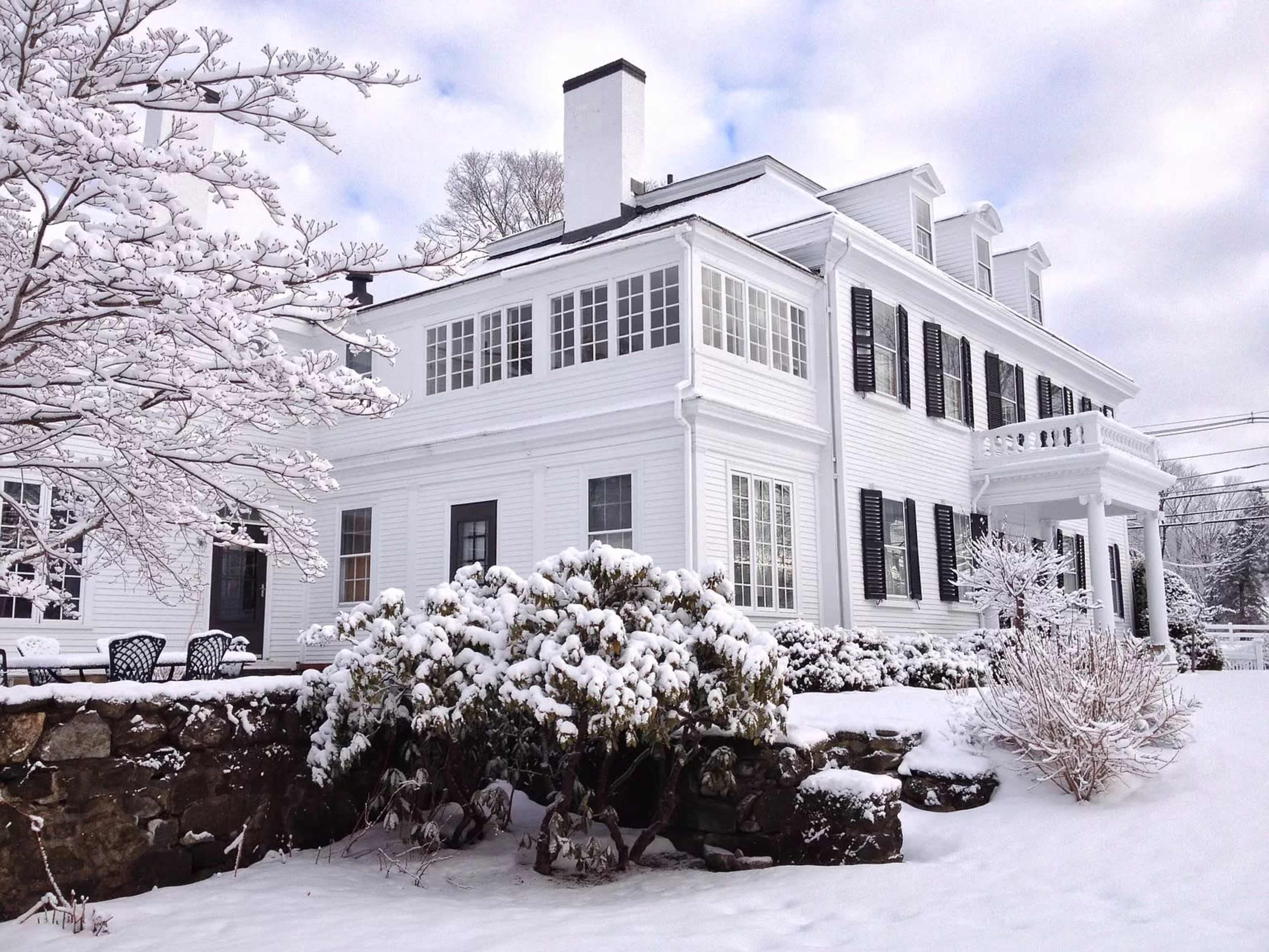 A large white house with multiple windows and a balcony is surrounded by snow-covered landscaping and trees.