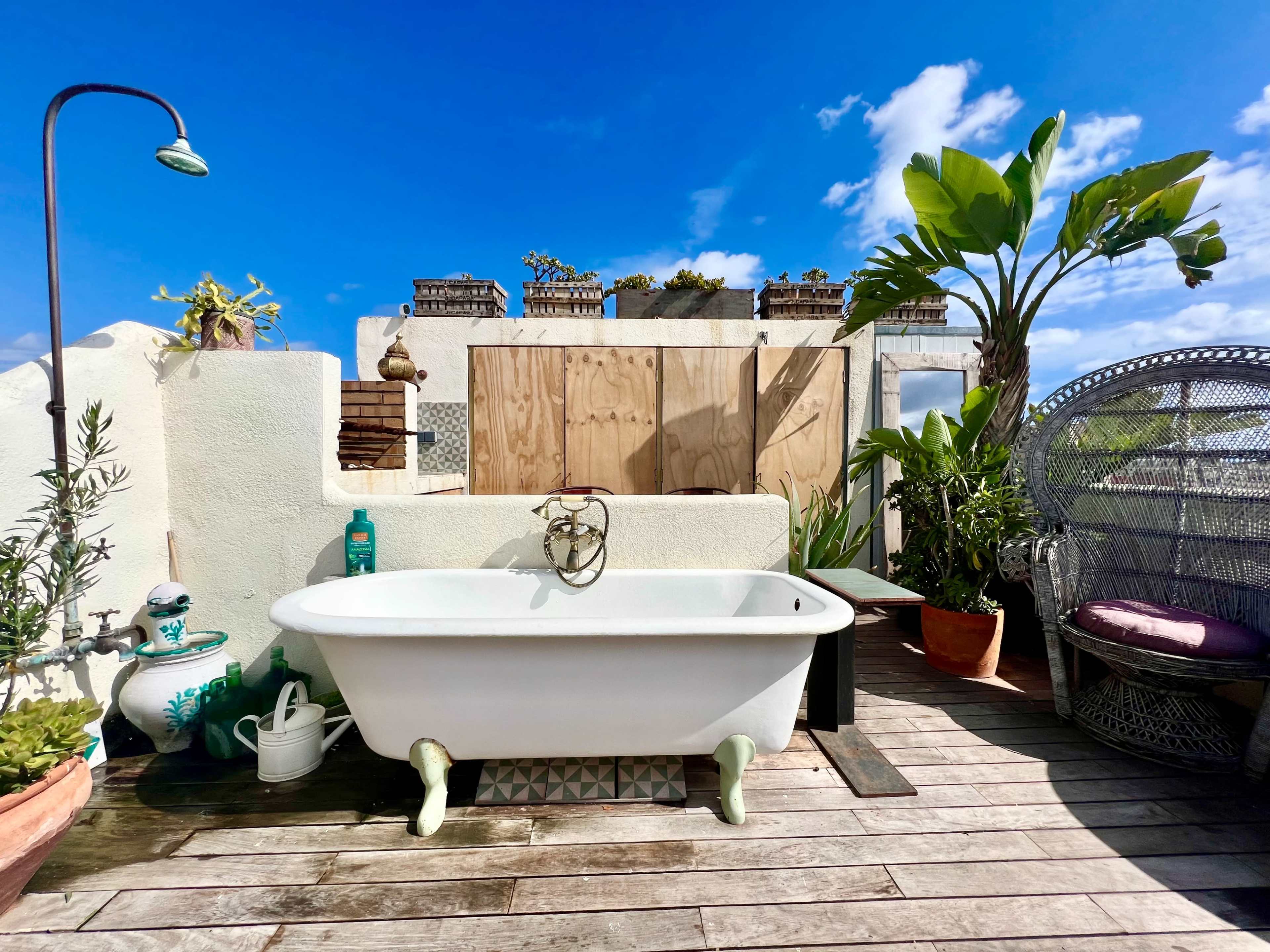 A vintage bathtub sits on a wooden deck surrounded by potted plants and a wicker chair under a blue sky with clouds.
