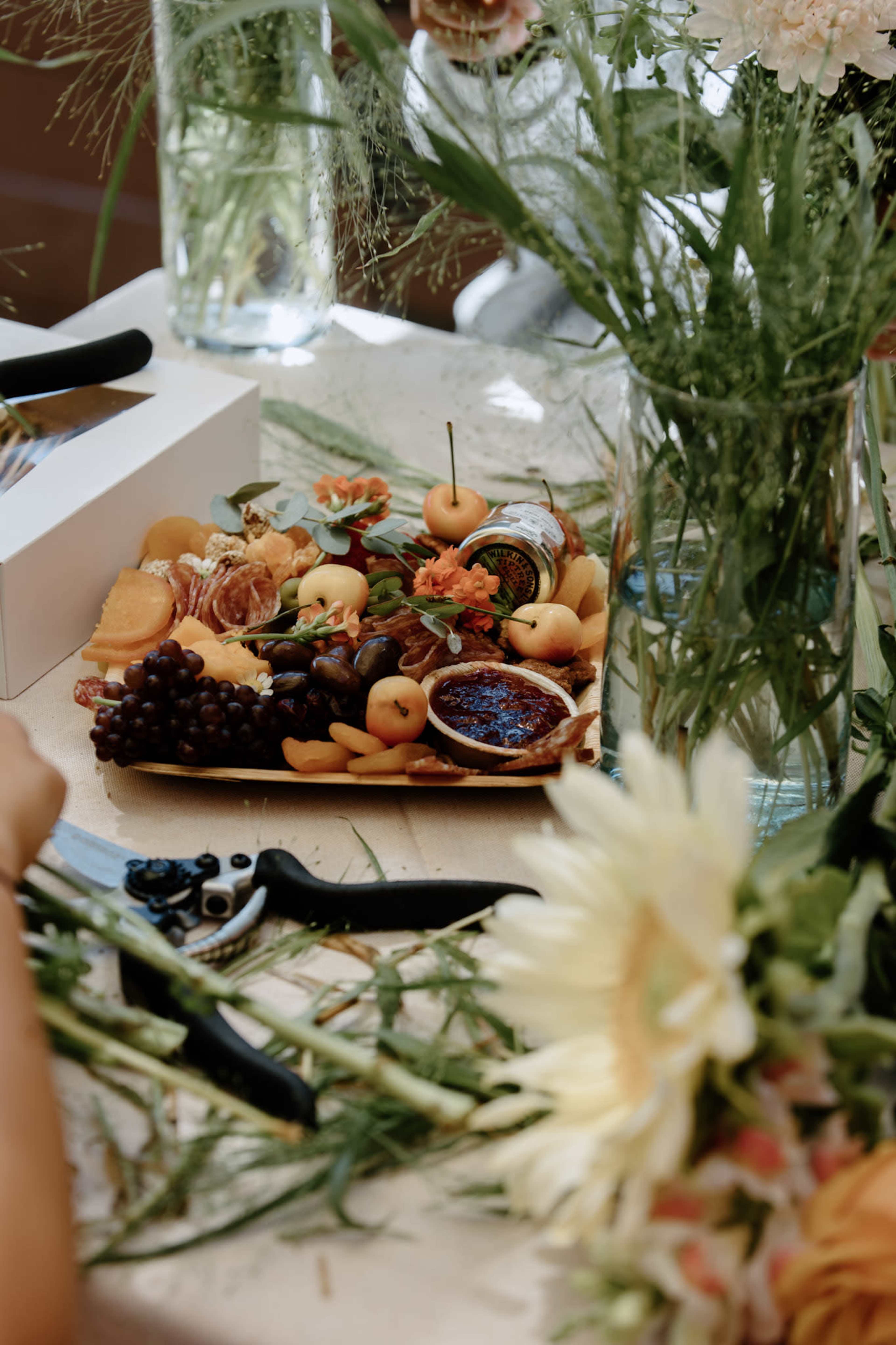A wooden platter filled with assorted fruits and decorative flowers is placed on a table beside gardening tools and vases.