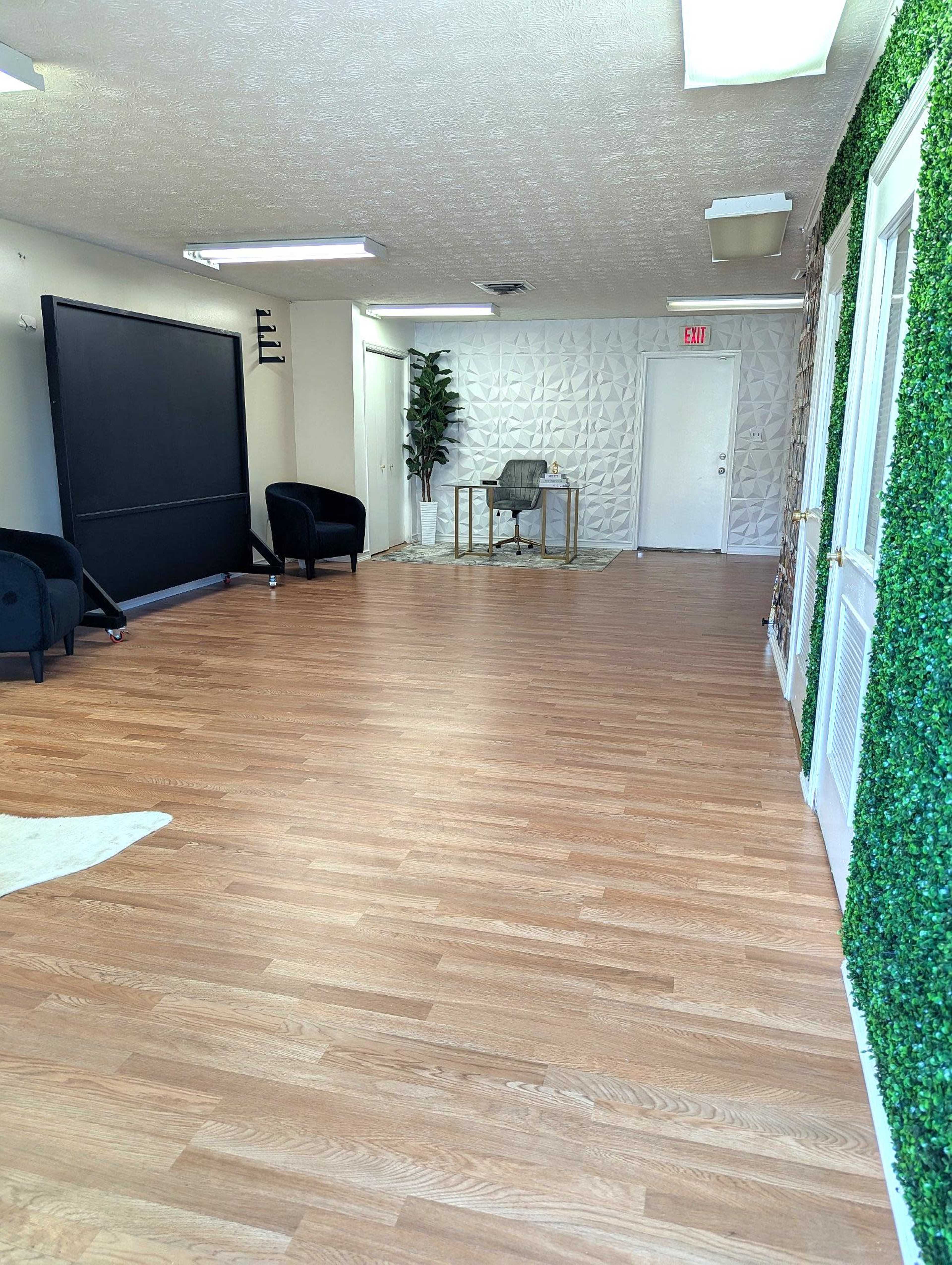The image shows a spacious room with wooden flooring, featuring a seating area with two black chairs, a desk at the far end, and a wall covered in a green plant decoration.