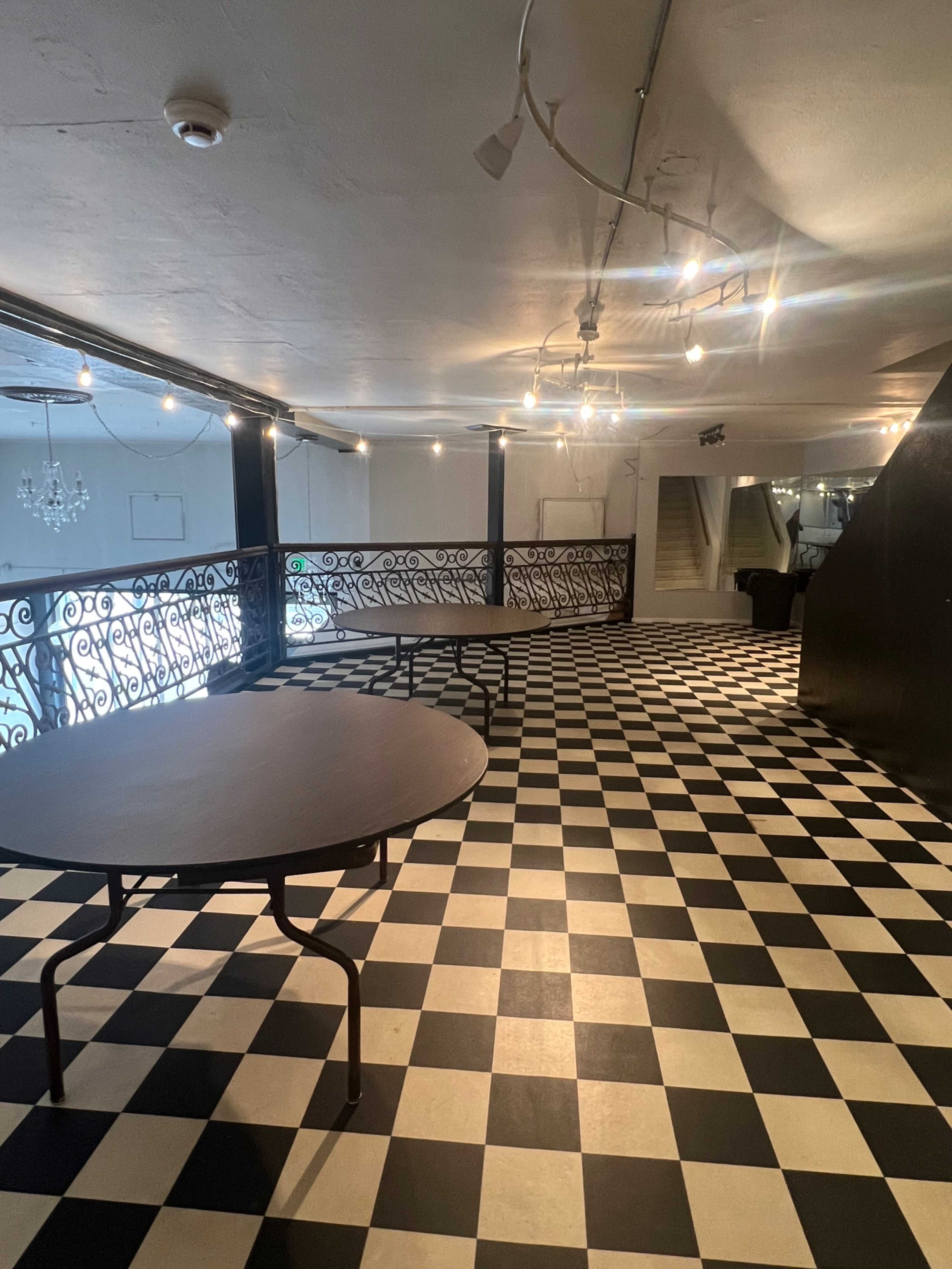 The image shows an indoor space featuring a checkered black and white floor with several round tables and decorative lighting overhead.