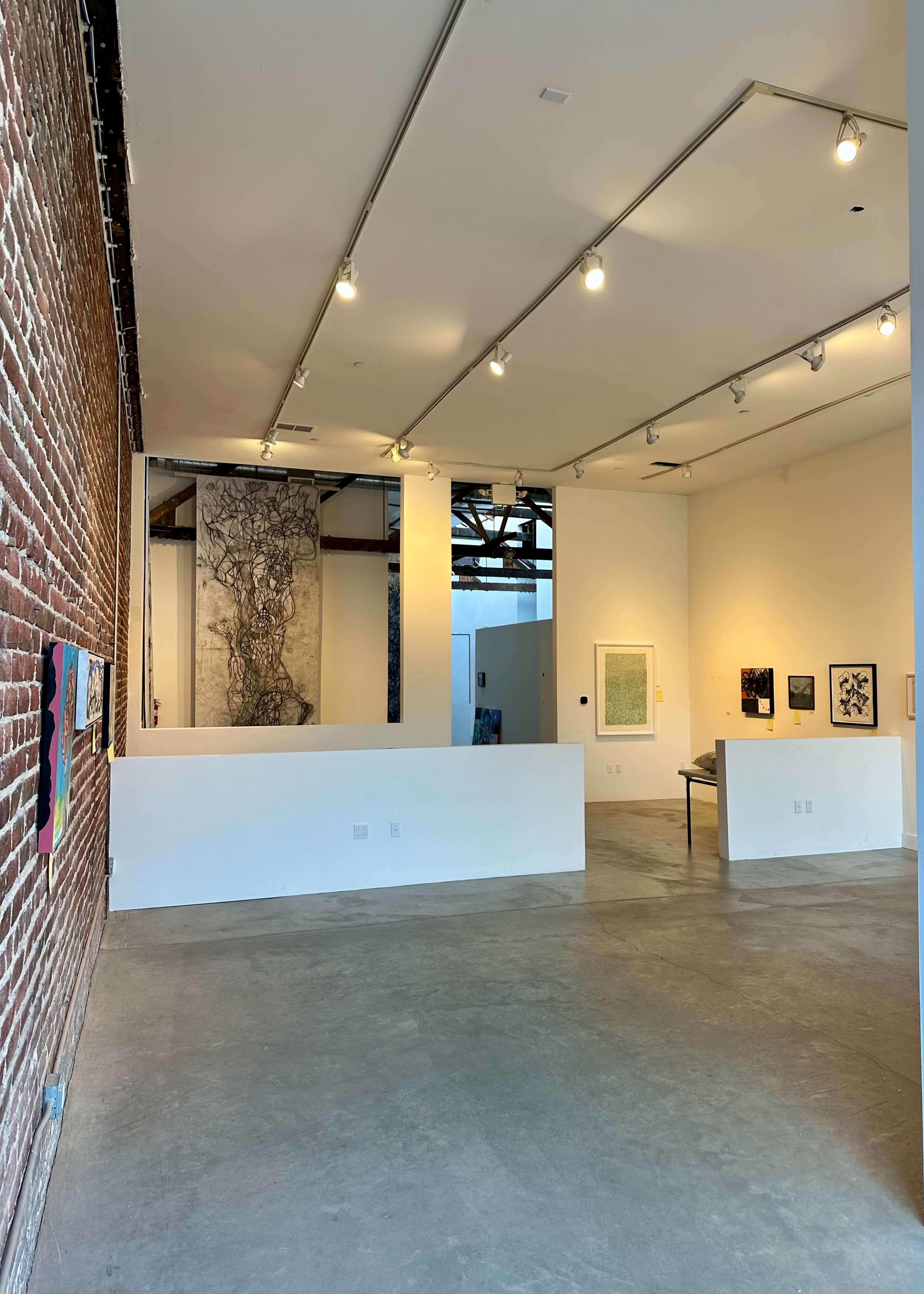 The image shows an art gallery featuring a mix of artworks on brick and white walls, with a concrete floor and track lighting illuminating the space.