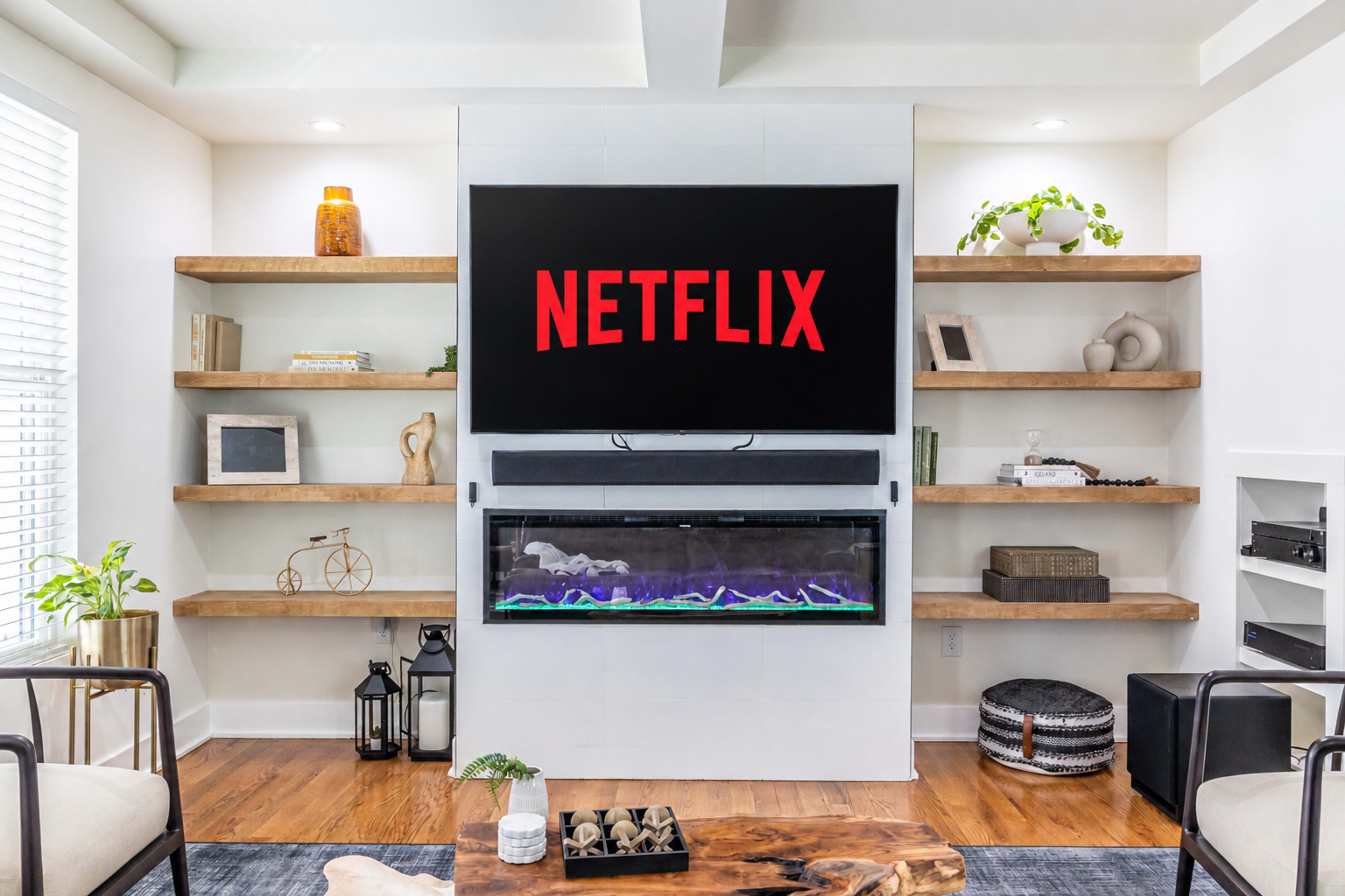 A modern living room features a television mounted above a sleek electric fireplace, flanked by wooden shelves displaying various decor items.