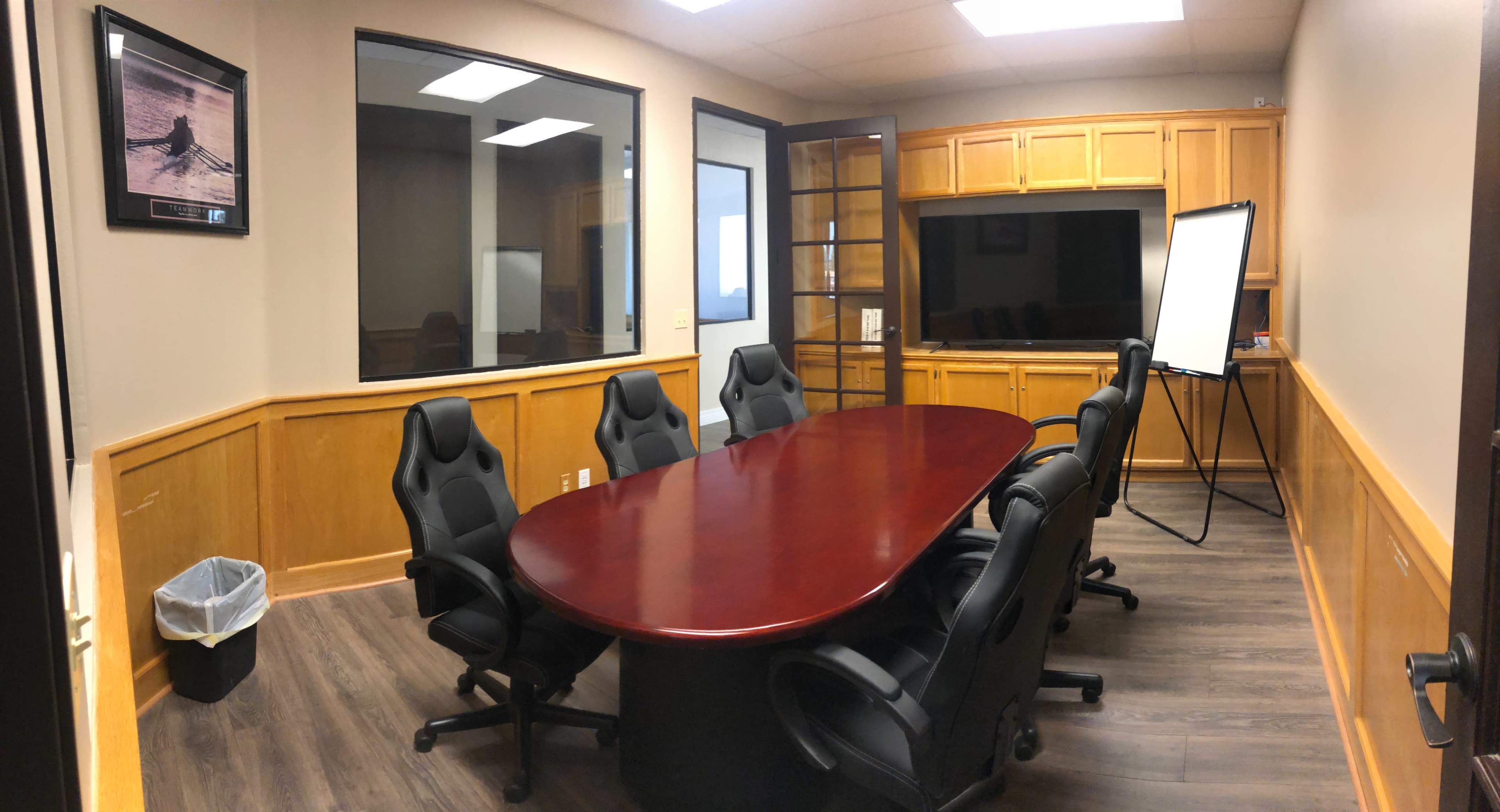The image shows a conference room with a large round table, several black office chairs, a whiteboard on a stand, and wooden cabinetry along the walls.