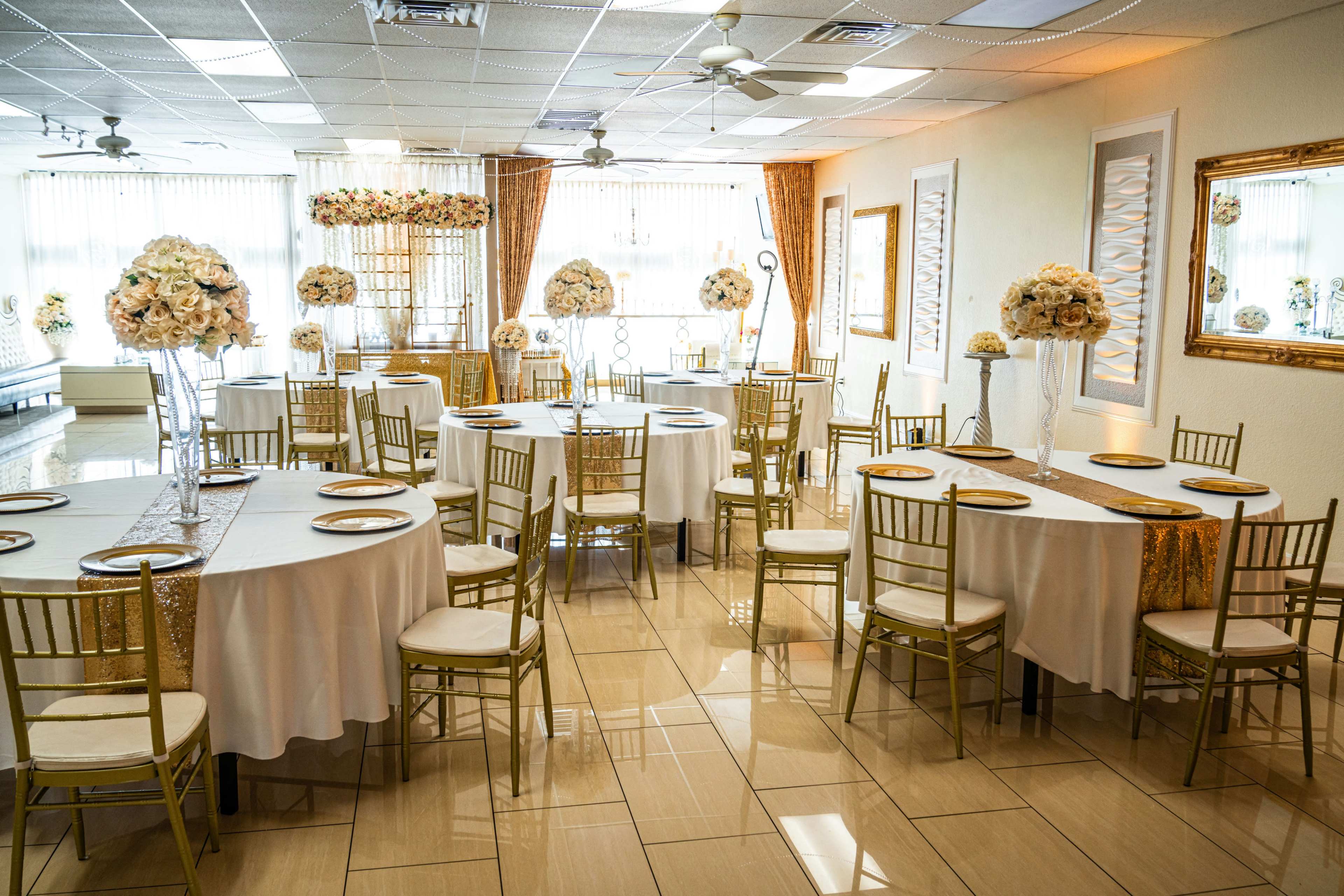 The image shows a banquet hall set up with round tables adorned with white tablecloths and gold-accented decorations, featuring floral centerpieces and elegant chairs.