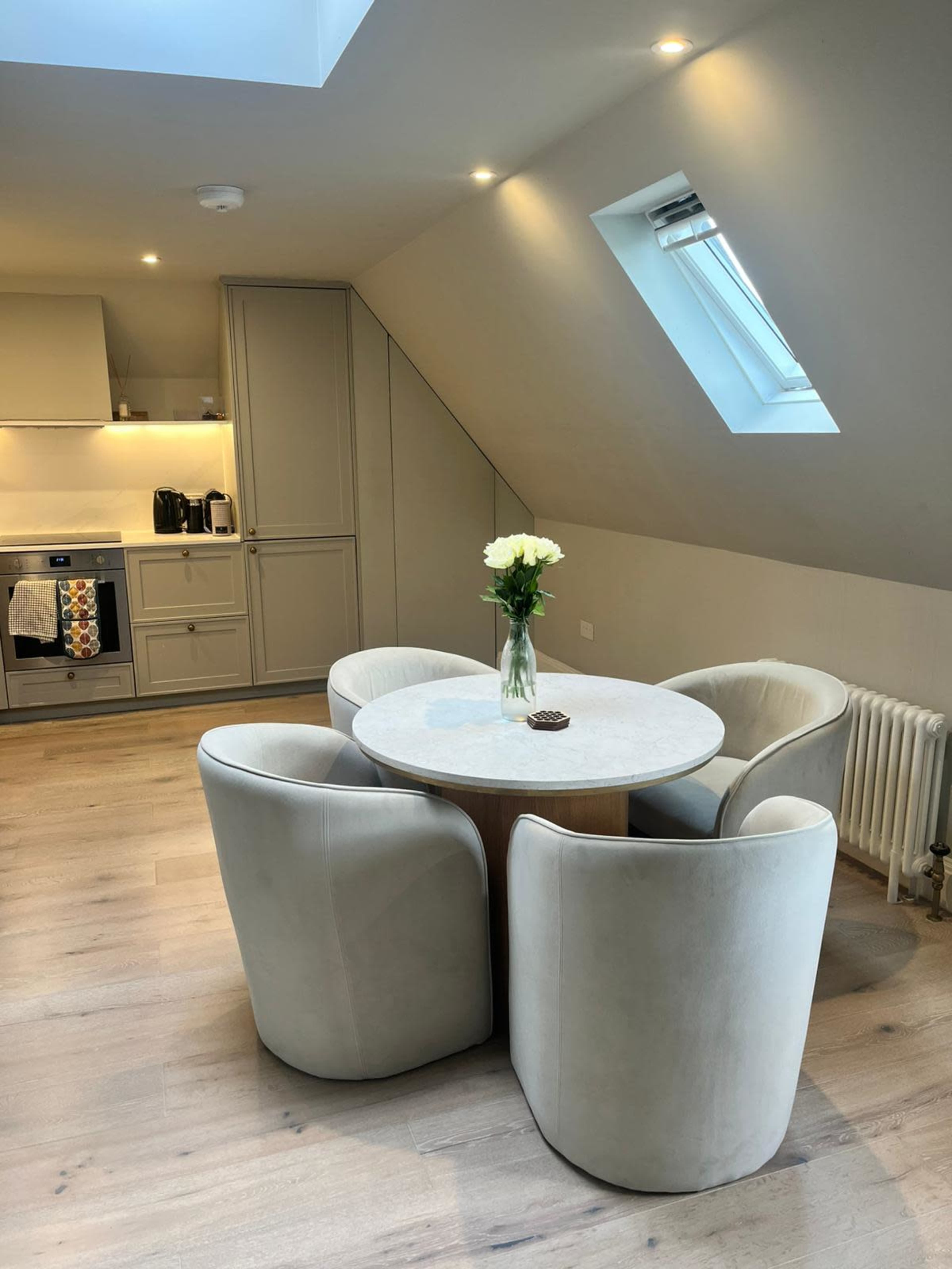 A small kitchen area features a round table surrounded by four upholstered chairs, with a vase of white flowers on the table.