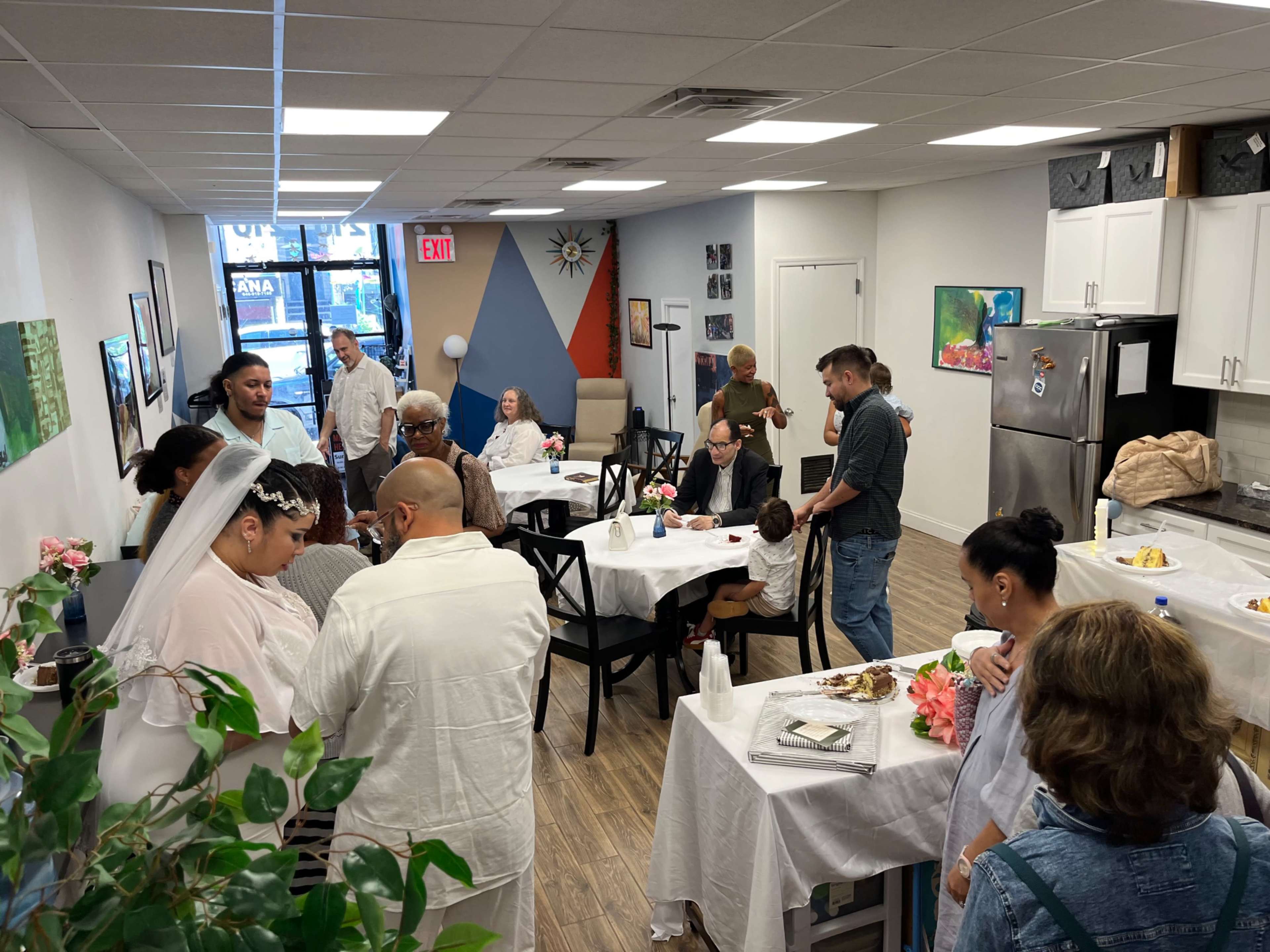 A group of people gather in a brightly lit room with tables set for a meal, engaged in conversation and enjoying each other's company.