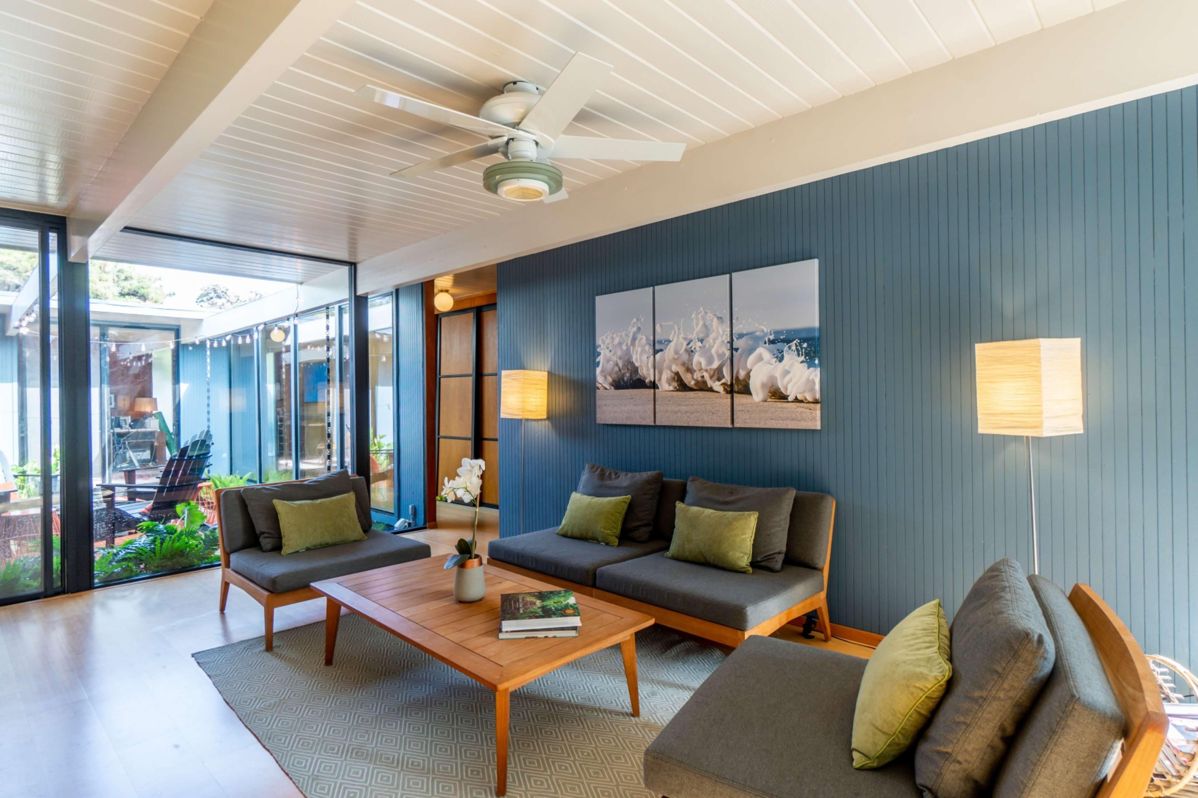 The image shows a modern living room featuring a wooden coffee table, gray sofas with green cushions, and a ceiling fan, with large windows overlooking a patio.