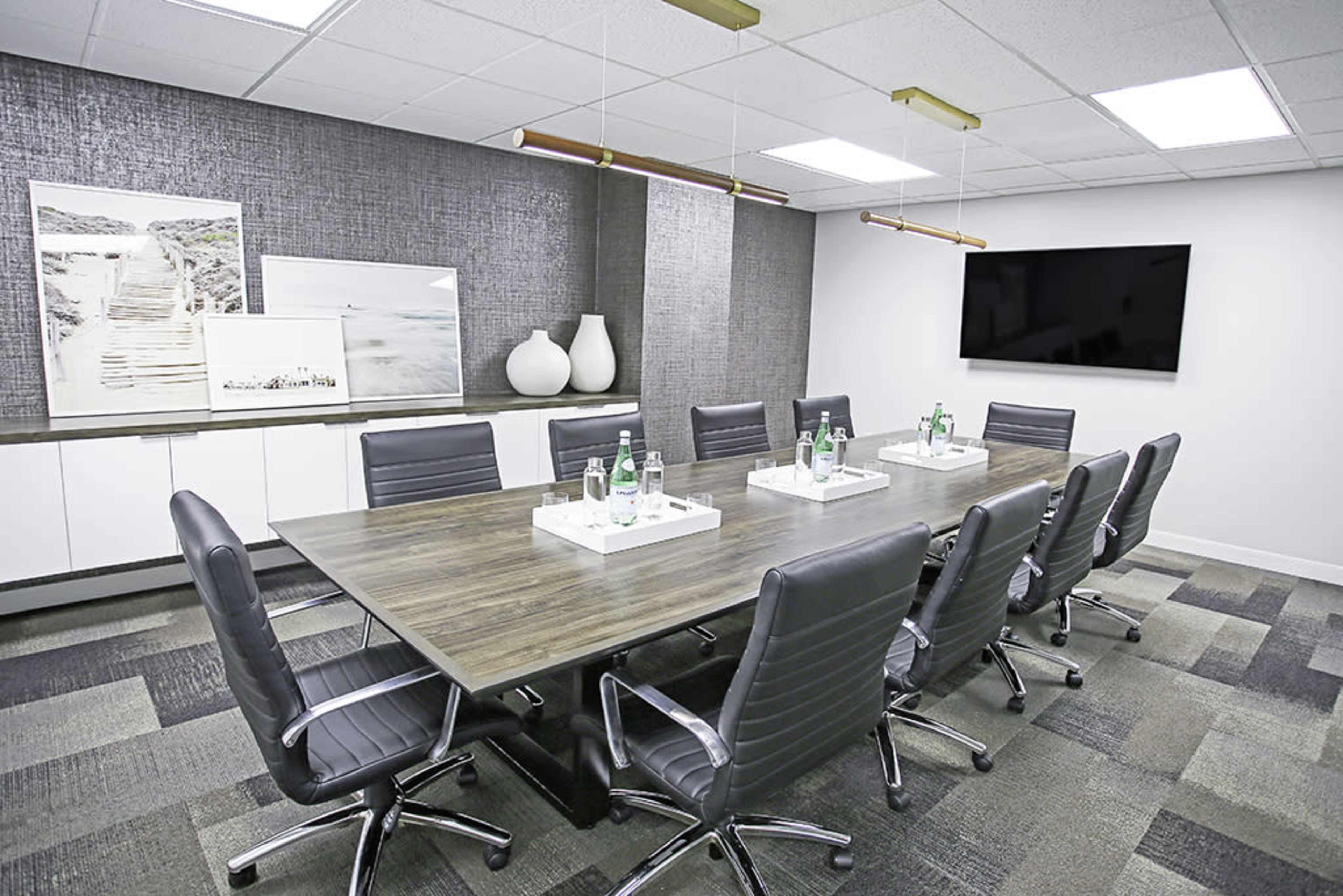 A modern conference room features a long wooden table surrounded by ergonomic chairs, with water bottles on white trays and wall-mounted artwork.