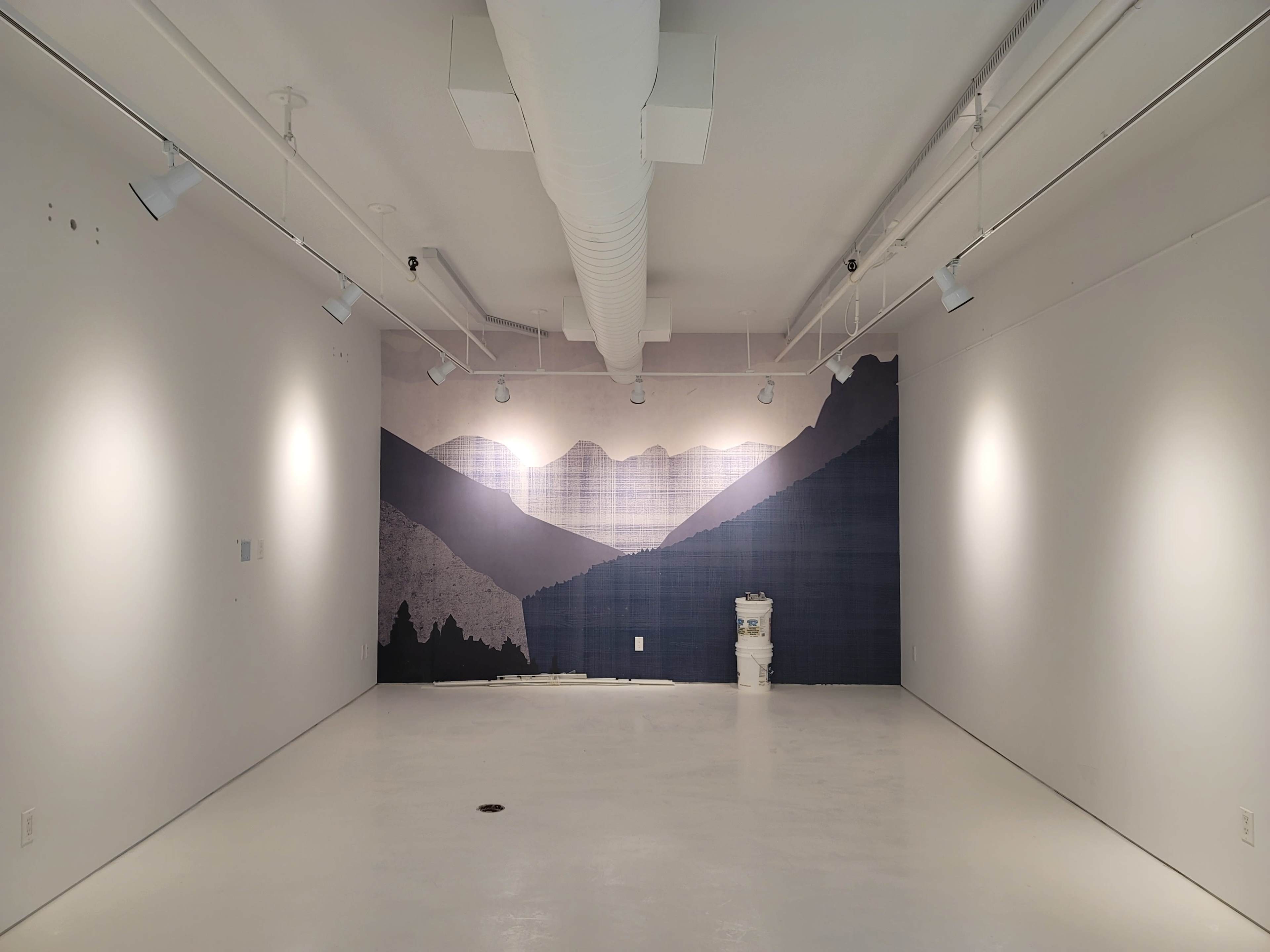 An empty gallery space features a large mural of mountains on one wall, with bright white walls and a concrete floor.