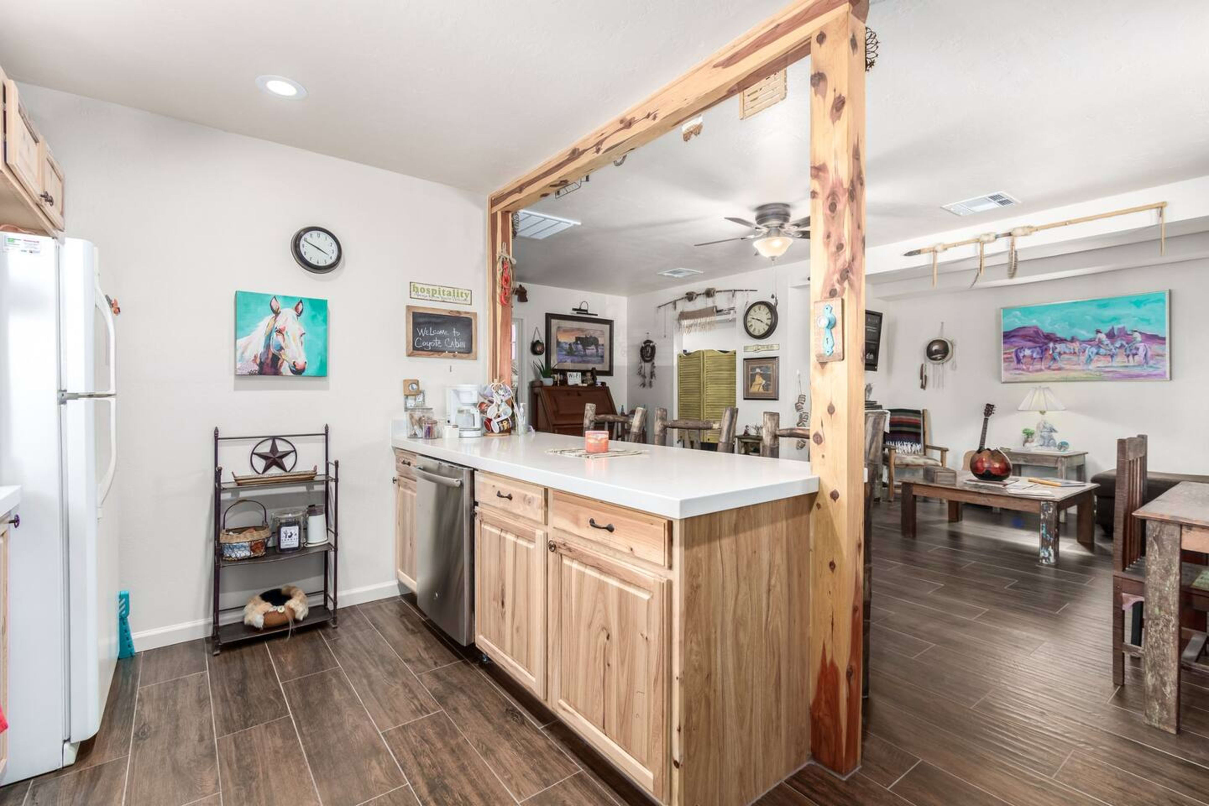 A modern kitchen with wooden cabinetry and a countertop dividing it from a dining area featuring rustic furniture and decorative artwork.