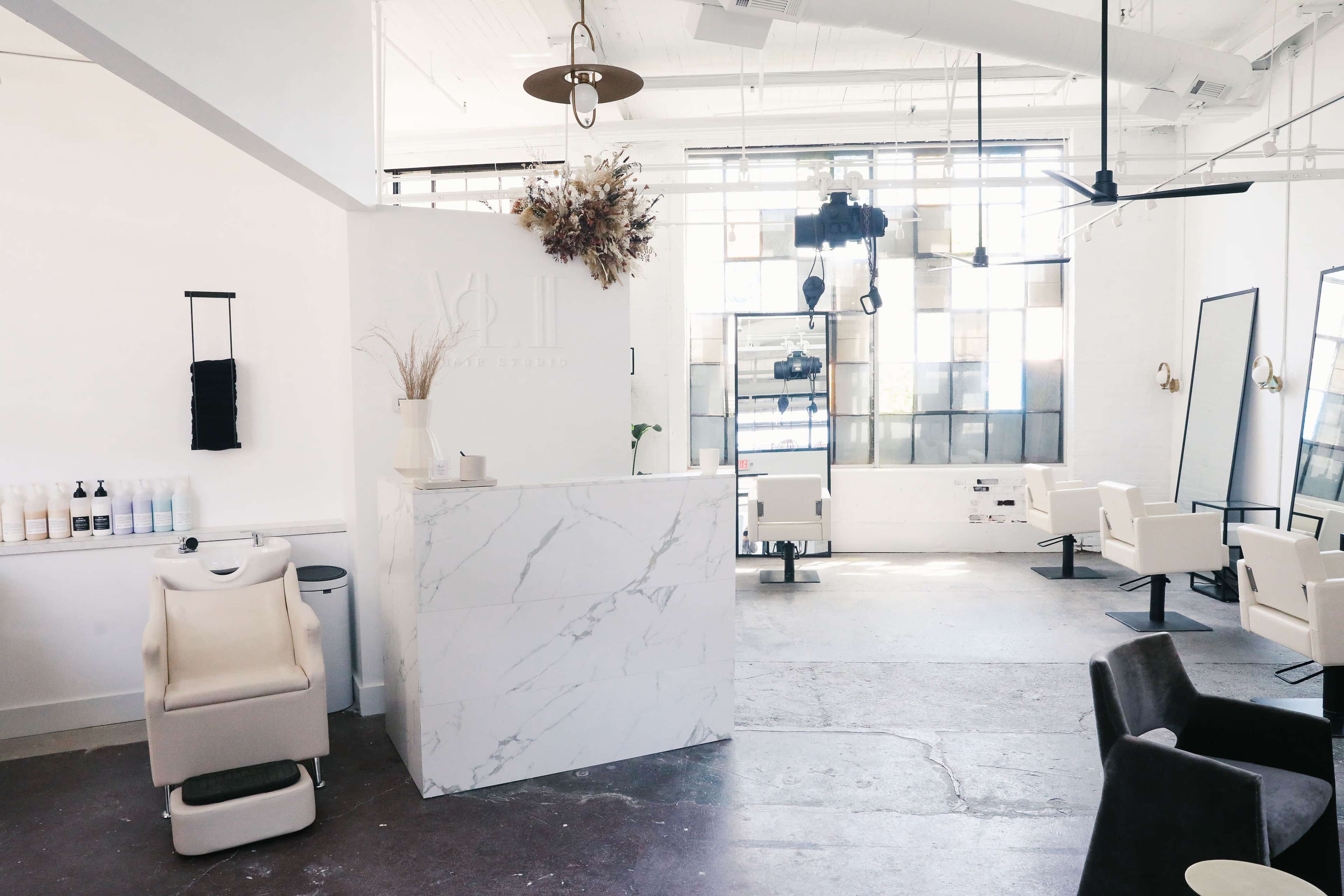 A modern hair salon with a marble reception desk, several styling chairs, large windows, and minimalist decor.