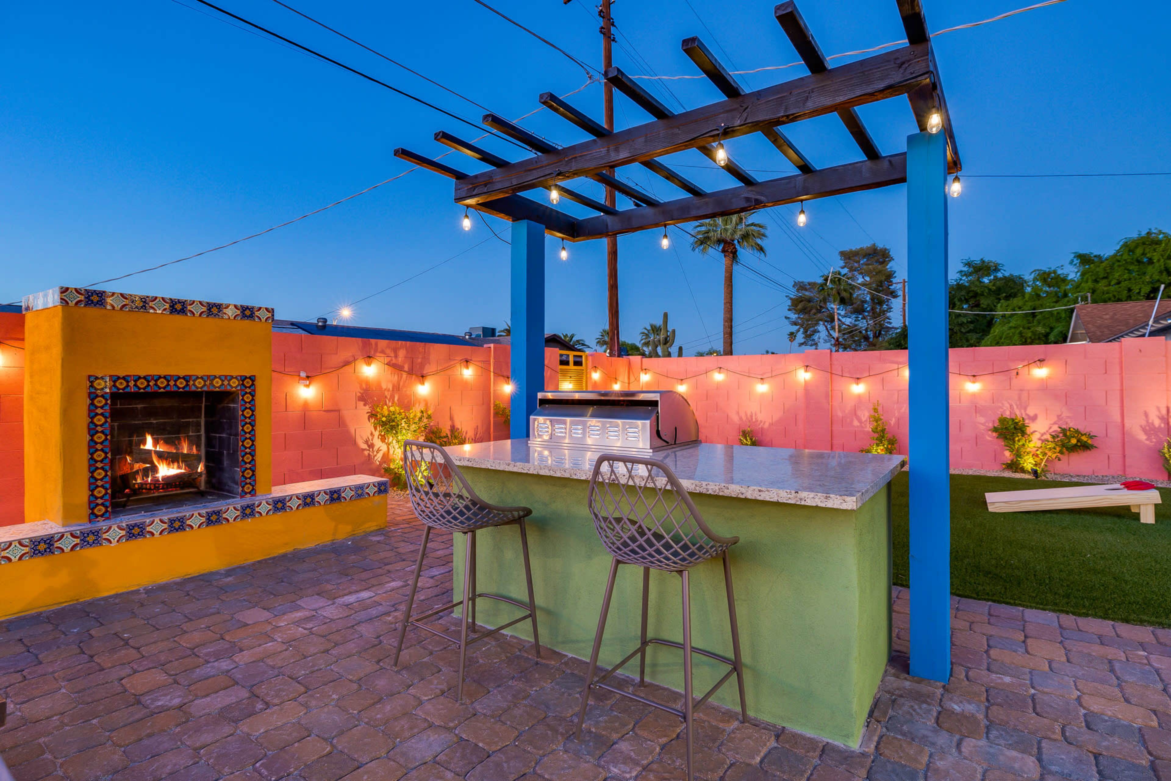 An outdoor patio with a modern bar area, a fireplace, and colorful walls, illuminated by string lights at dusk.