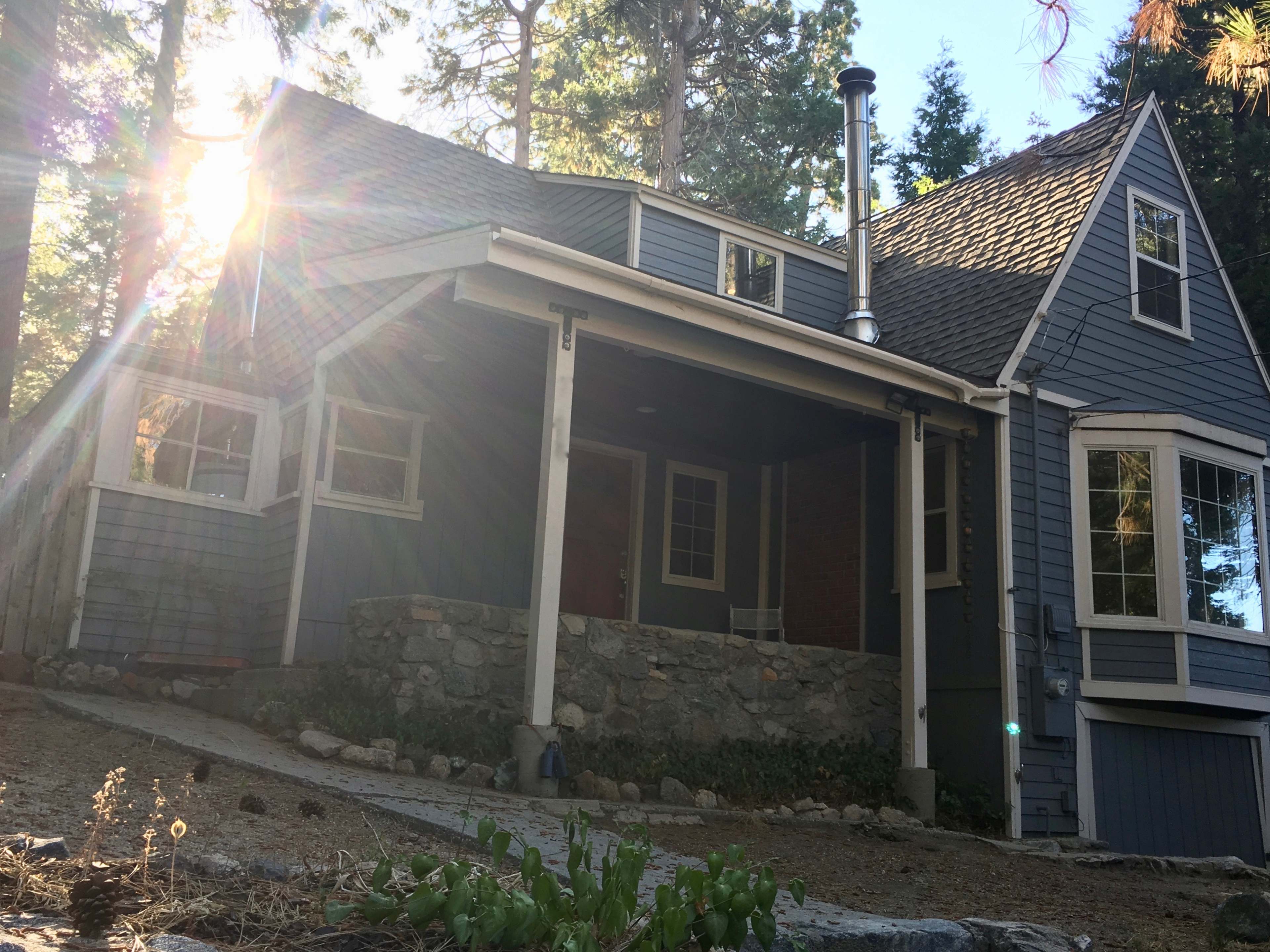A two-story blue house with a stone foundation and a chimney is surrounded by trees.