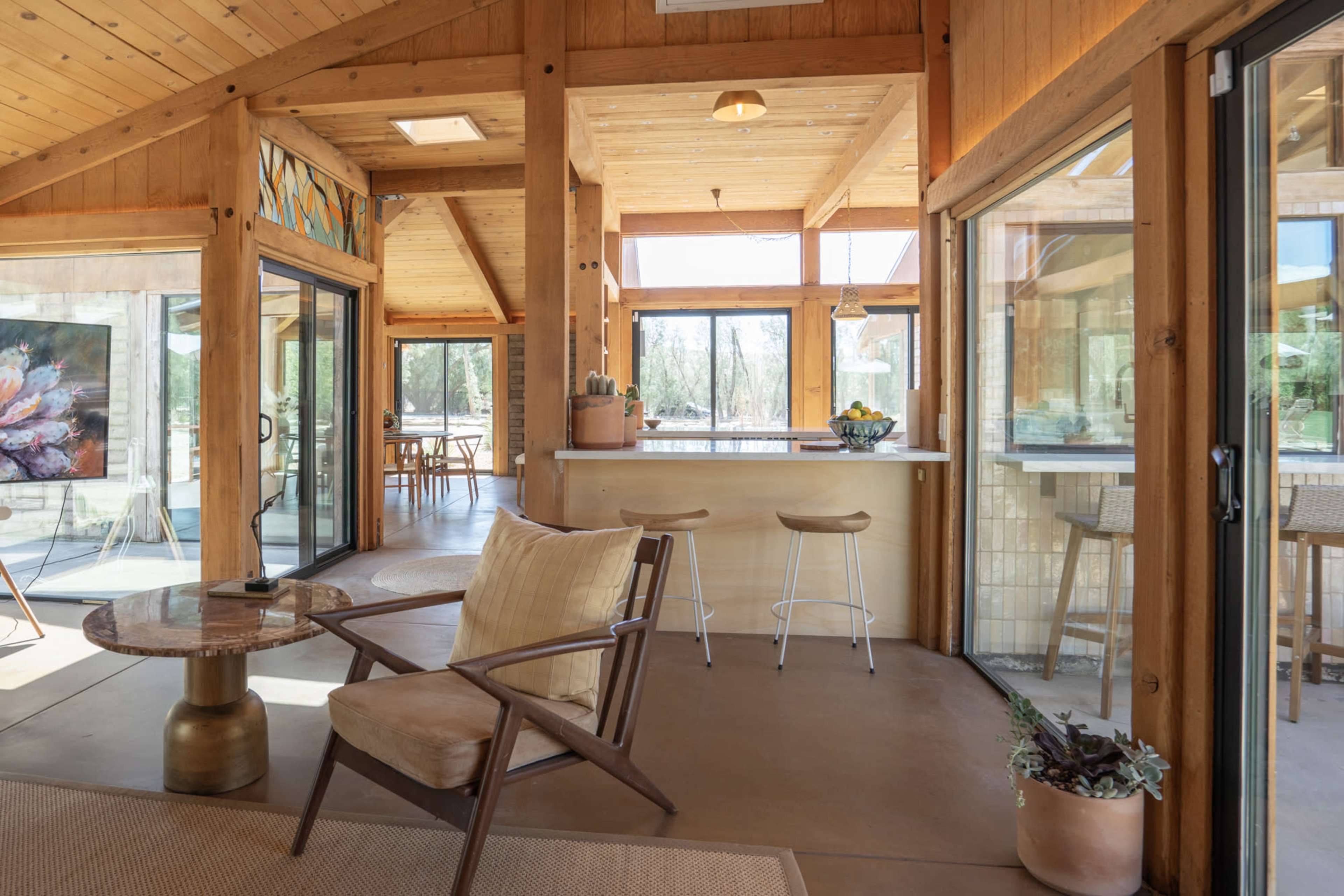 The image shows a modern, brightly lit interior of a home with wooden beams, large windows, and a minimalist living area featuring a chair and a round table.