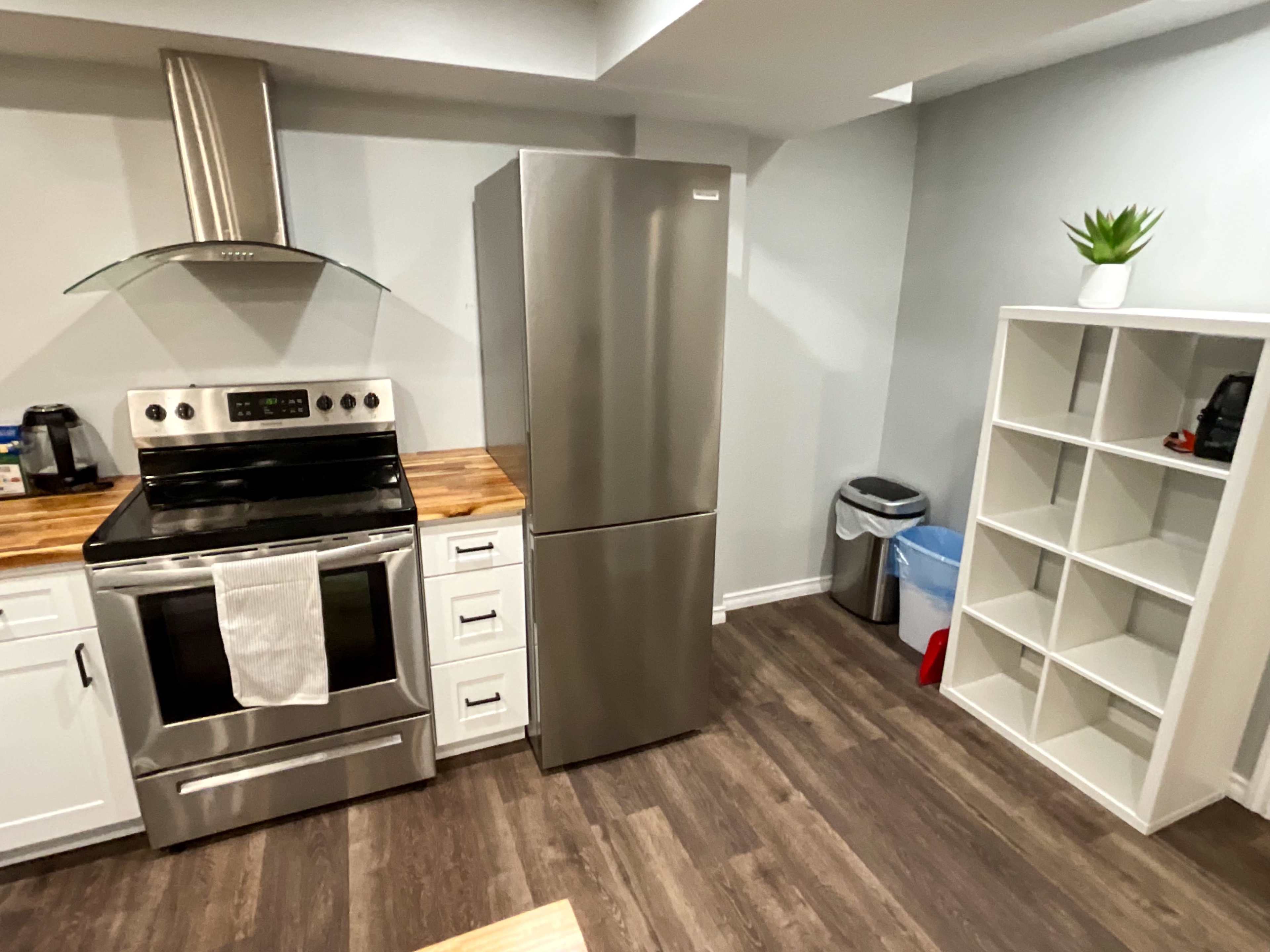 The image shows a modern kitchen with stainless steel appliances, including a refrigerator and an oven, a wooden countertop, and a white shelf unit against the wall.