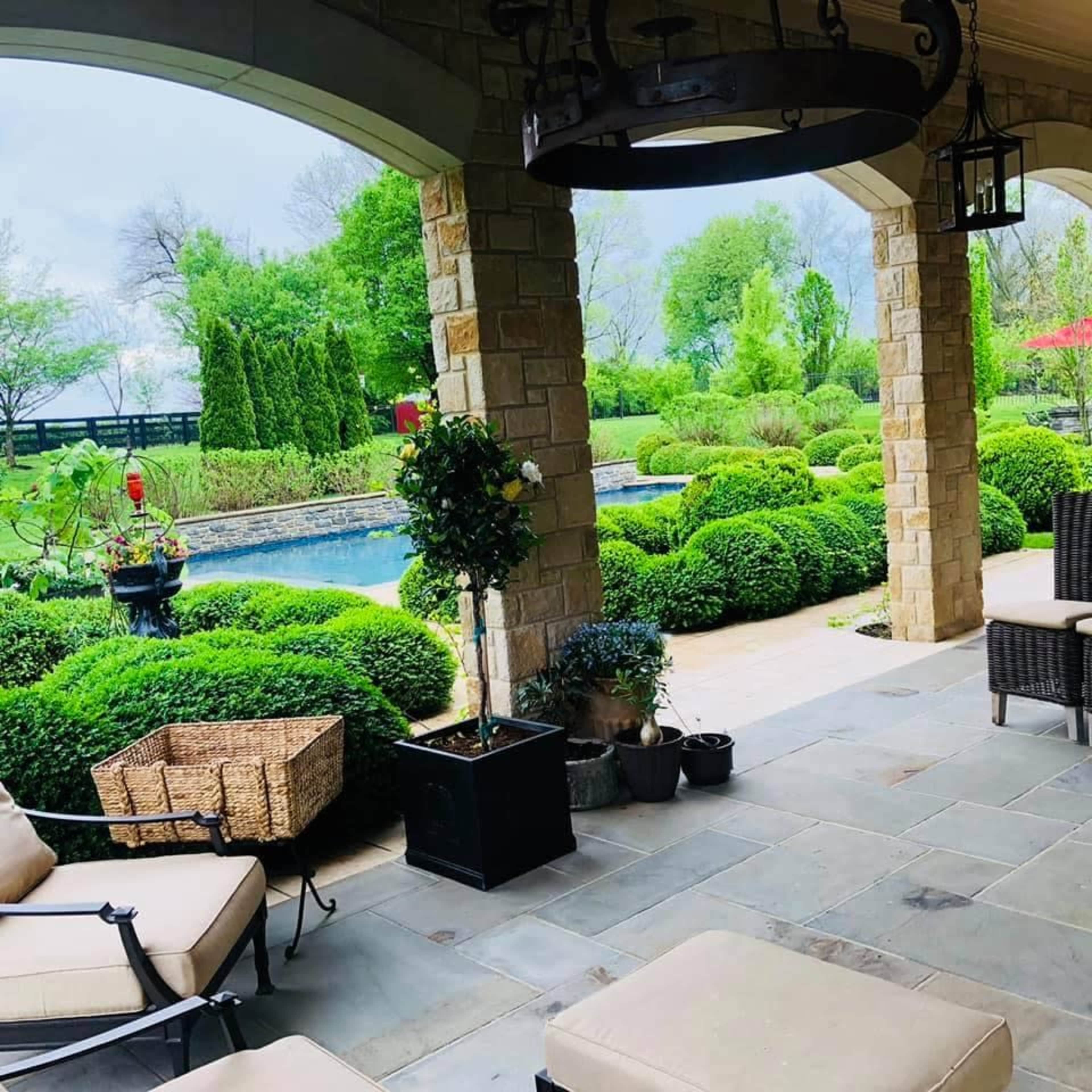 A covered patio area with stone columns overlooking a landscaped yard featuring a swimming pool and neatly trimmed hedges.