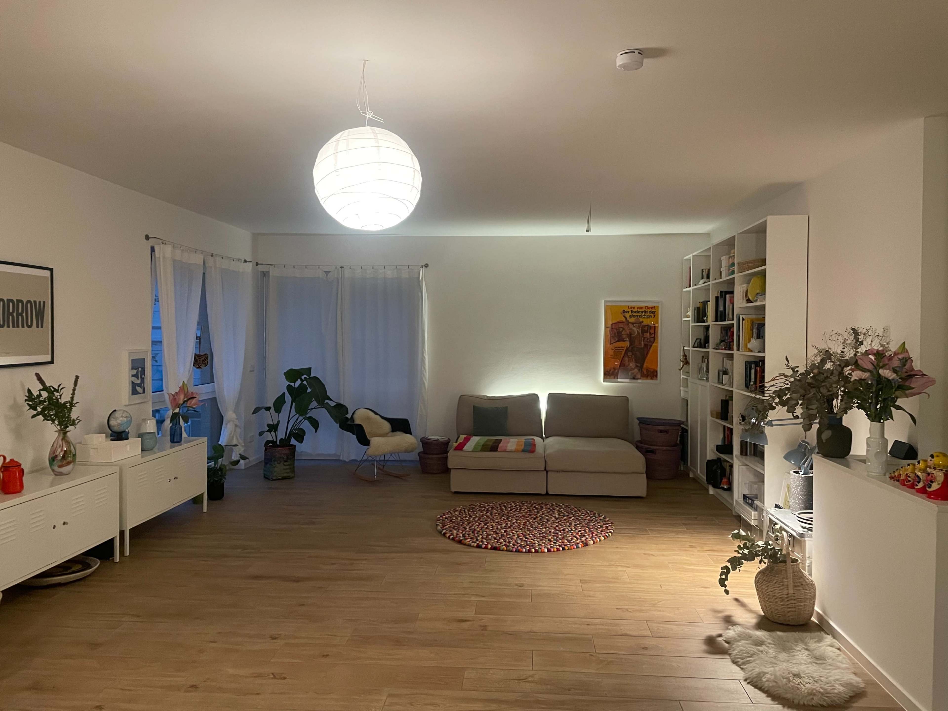 A spacious, minimalistic living room with a large window, a light fixture, a couch, a bookshelf, a circular rug, and decorative plants.