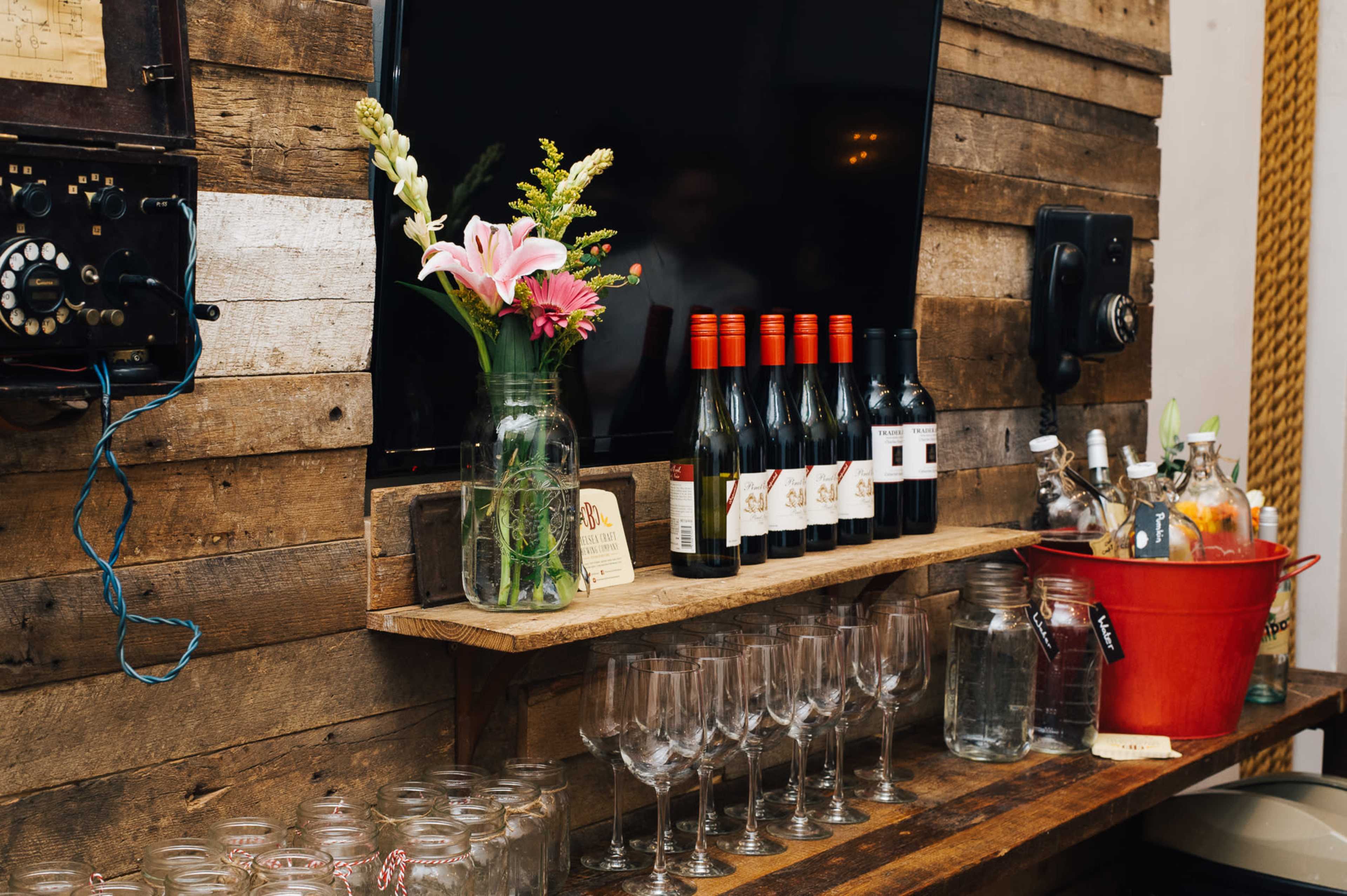 A rustic wooden bar setup features a shelf with wine bottles, a floral arrangement, glasses, and a red bucket.