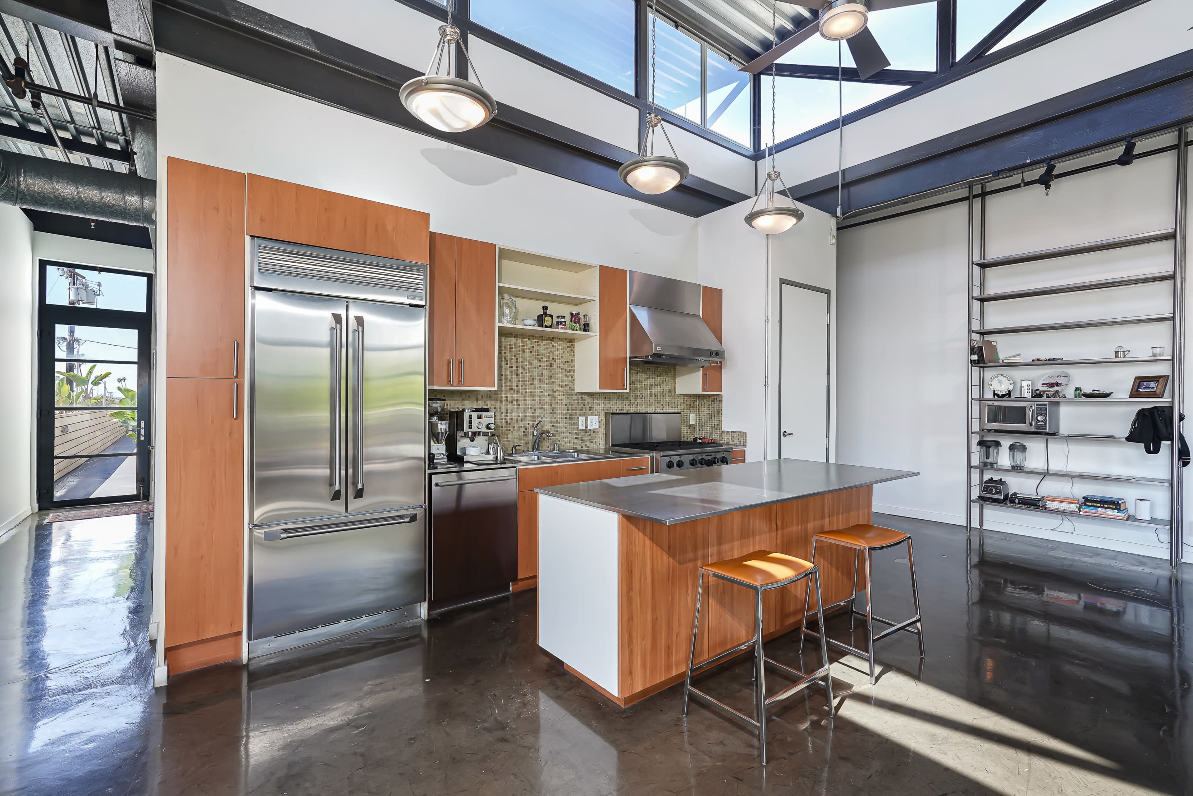 The image shows a modern kitchen with stainless steel appliances, wooden cabinetry, and a central island with two stools.