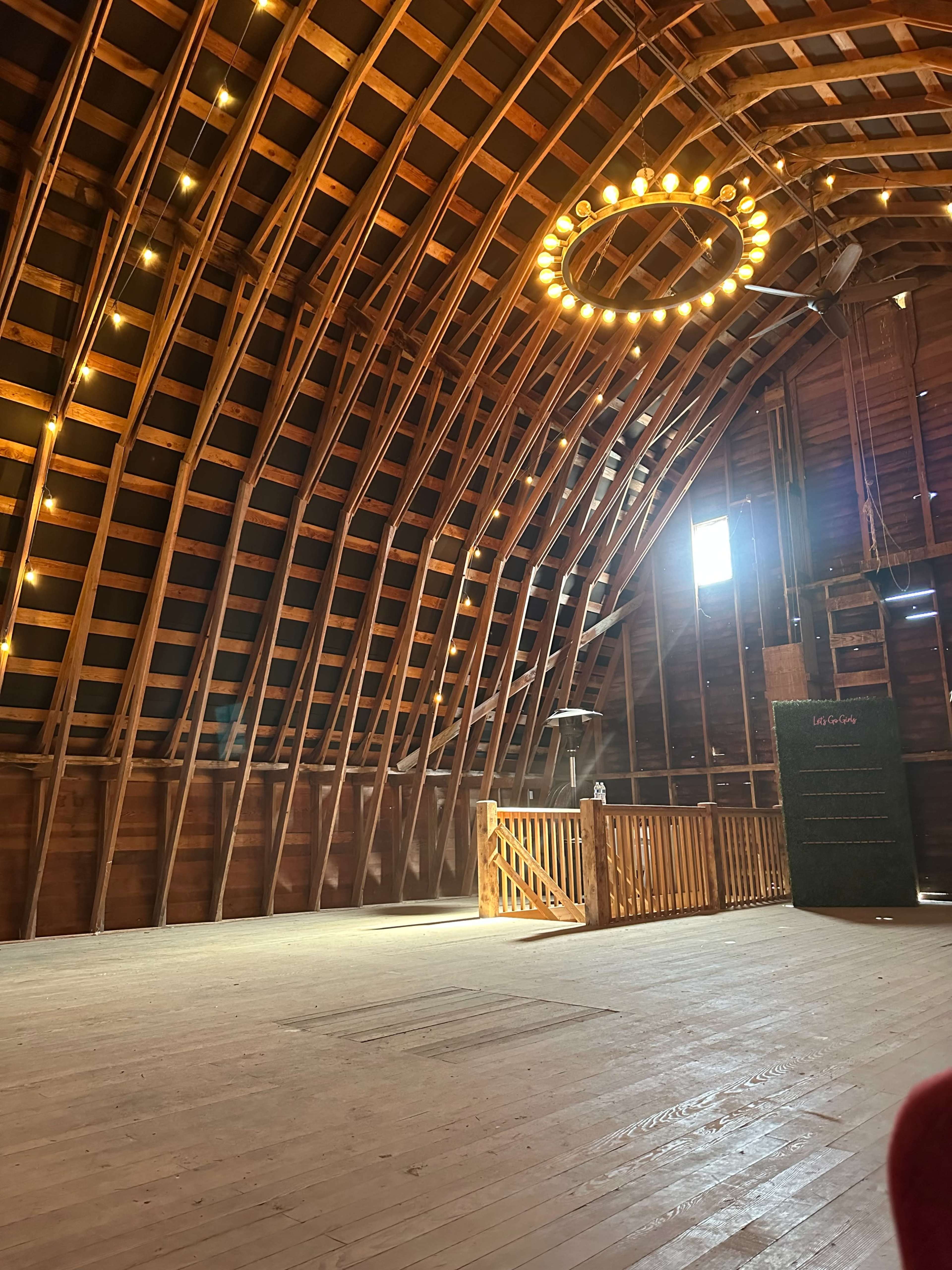 The image shows the interior of a wooden barn featuring arched wooden beams, a circular chandelier with lights, and a wooden stage area at one end.