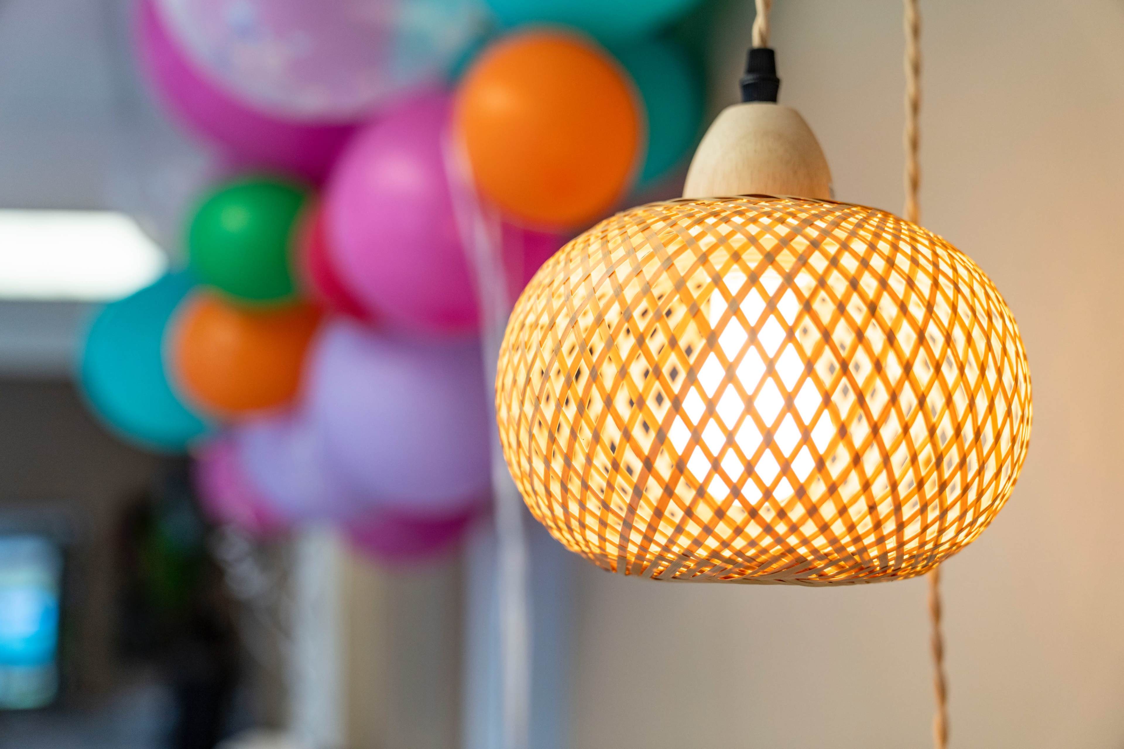 A woven pendant lamp is illuminated in front of a cluster of colorful balloons.
