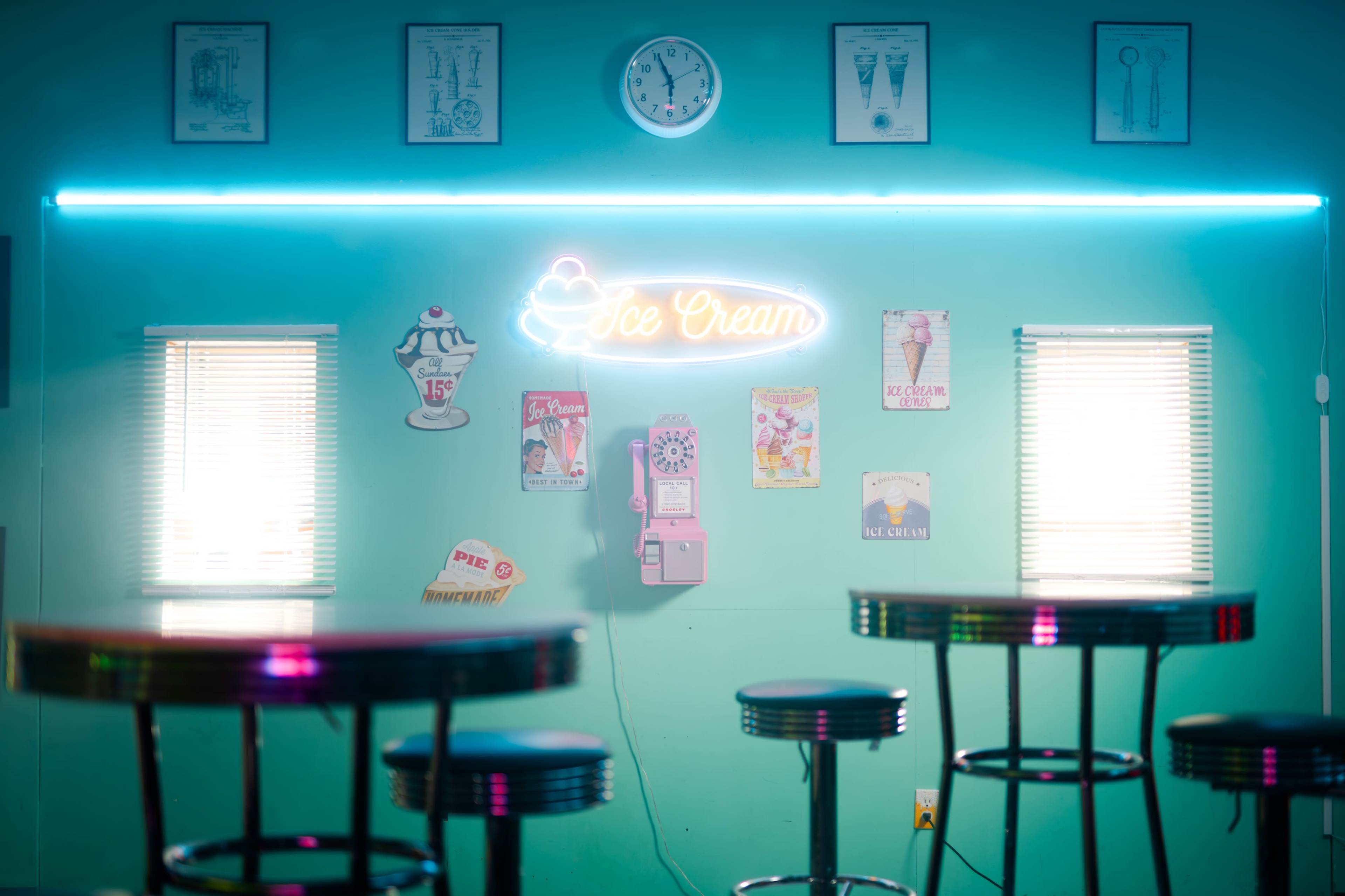 The image shows a brightly colored ice cream shop interior with a neon "Ice Cream" sign, a clock on the wall, and various vintage posters displayed above round tables with chrome stools.