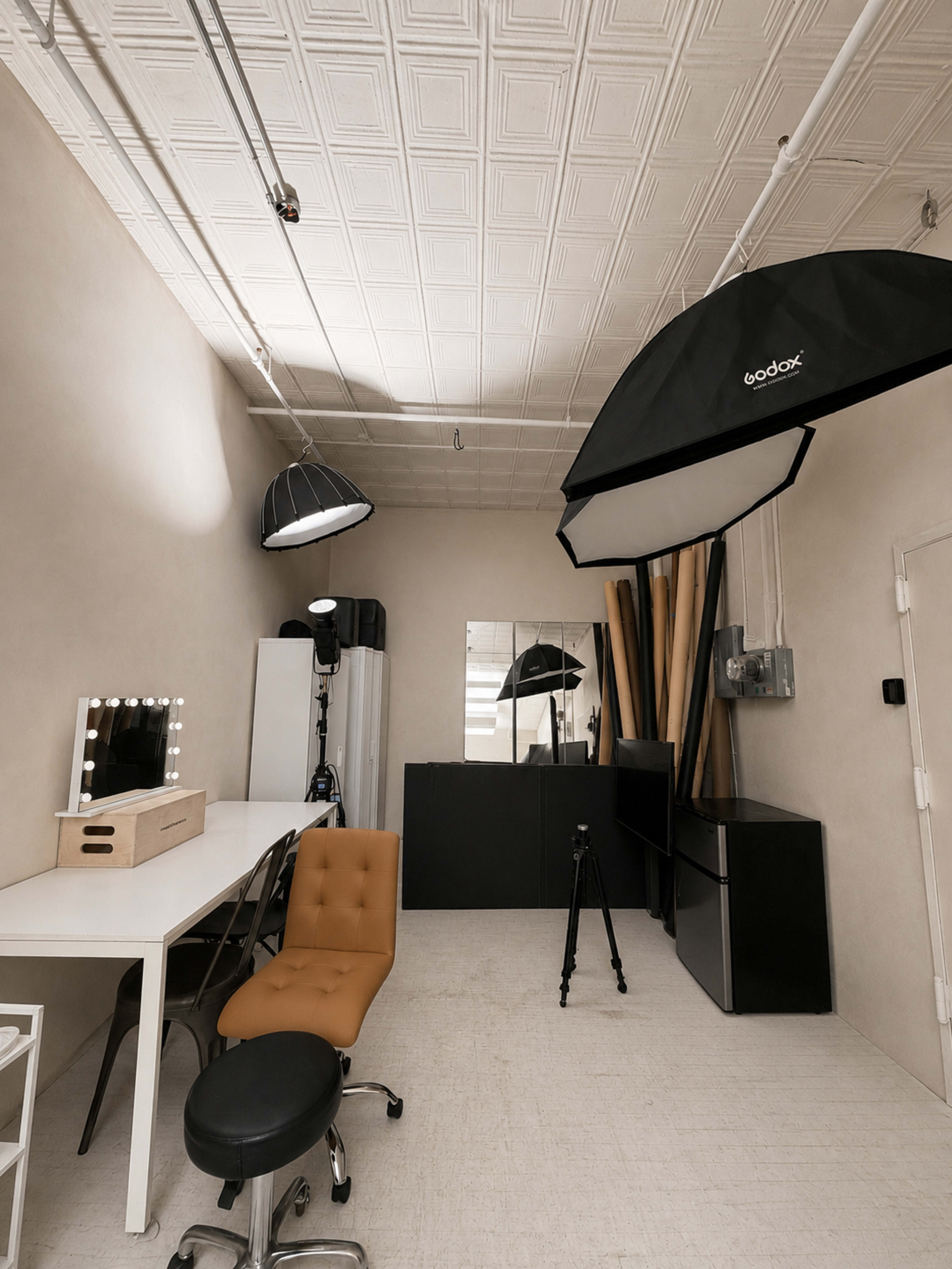 The image shows a well-organized photography studio featuring a large softbox, a makeup vanity with lights, a black backdrop, and various photography equipment.