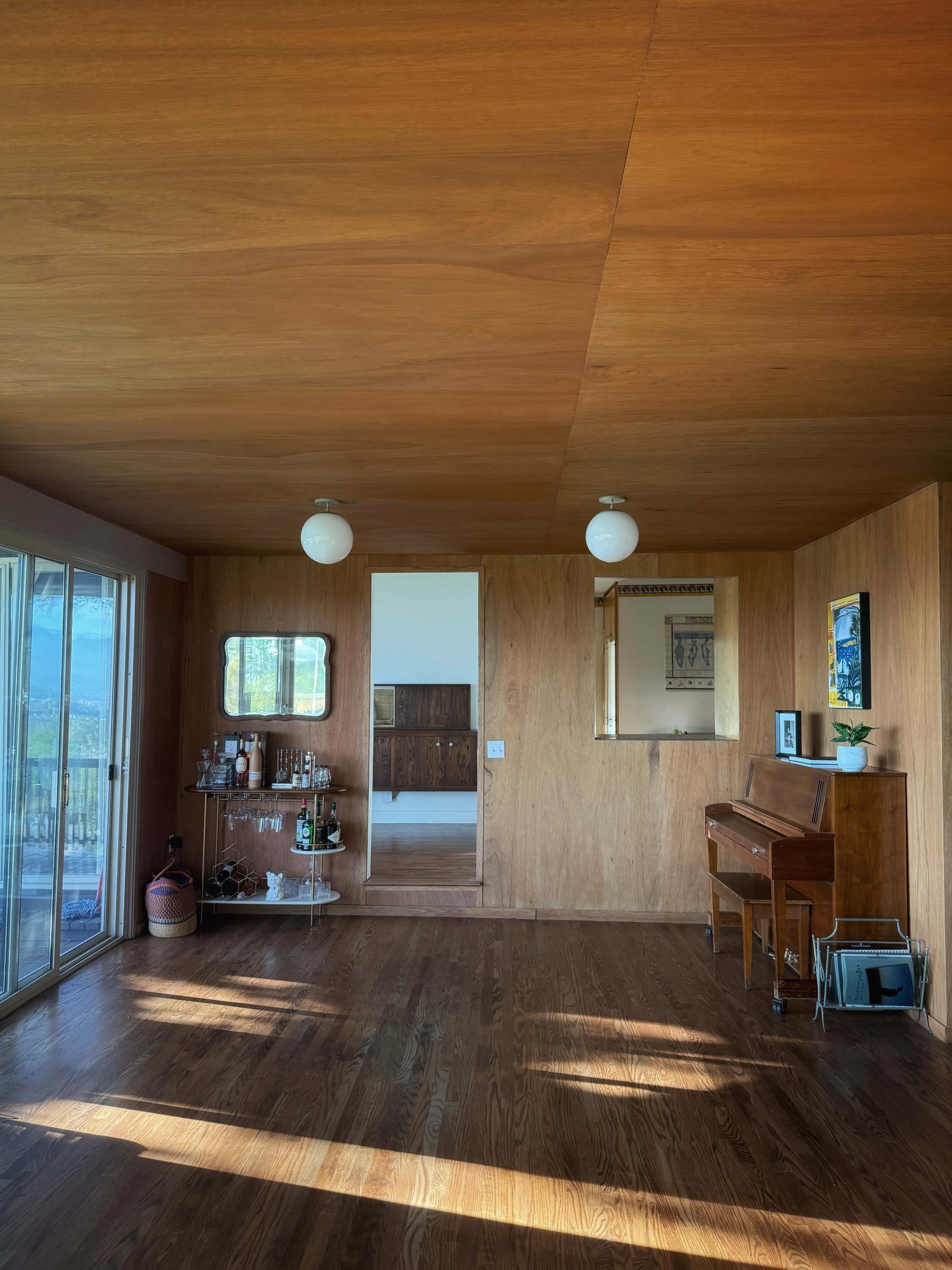 The image shows a wooden-paneled interior room featuring a piano, a bar cart, and large glass doors leading to an outdoor area.