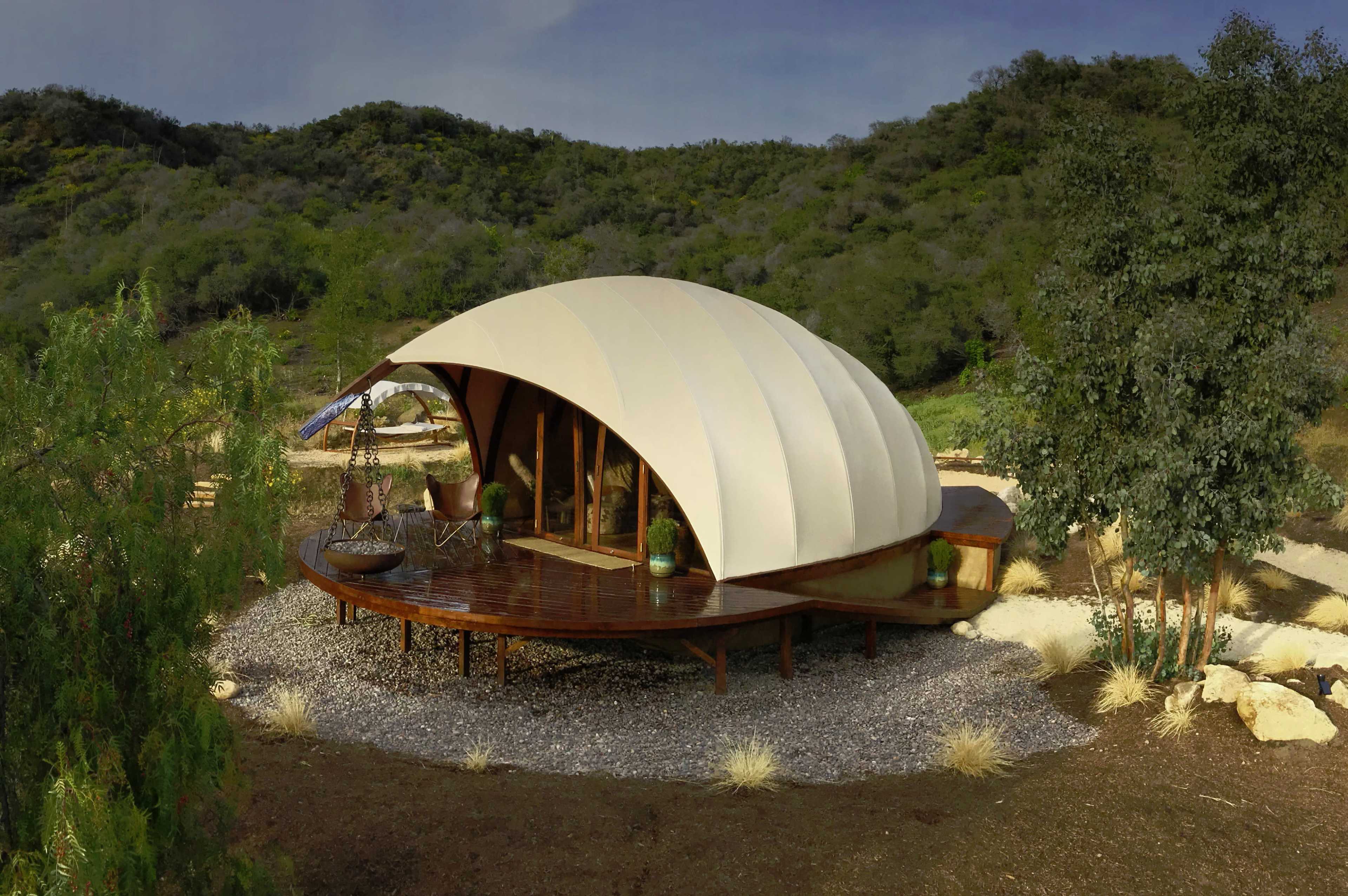 The image shows a modern, dome-shaped structure with a curved white roof situated on a wooden deck surrounded by greenery and gravel landscaping.