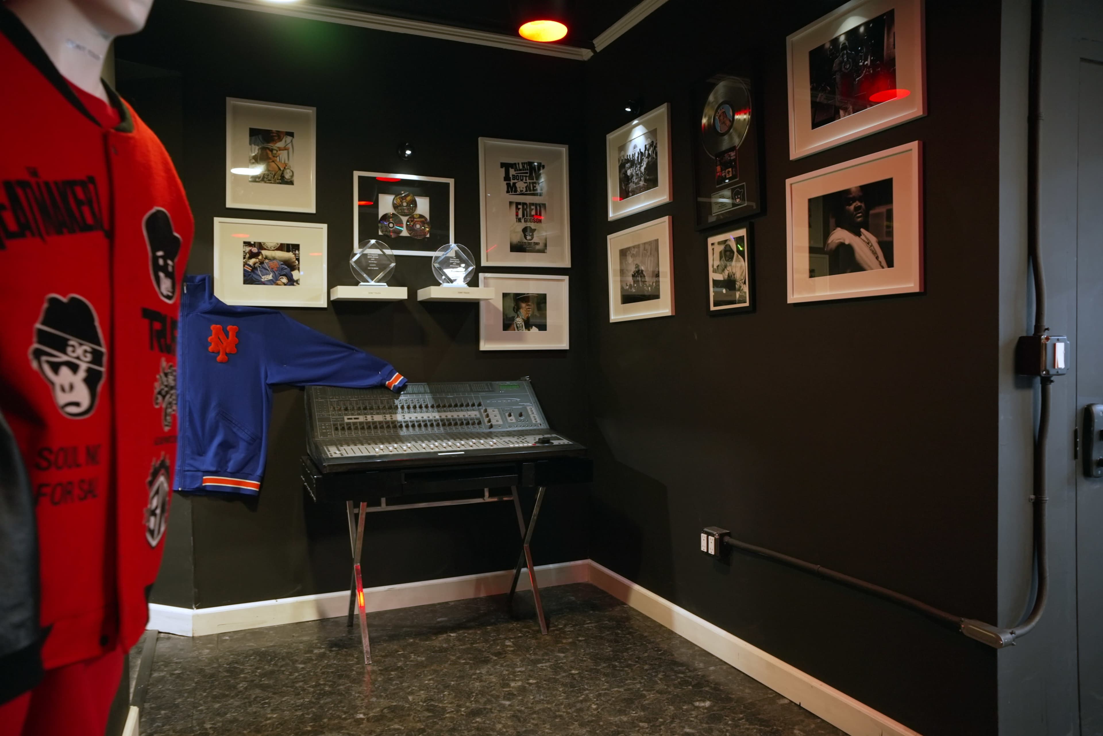 A corner of a music studio featuring a mixing console, framed photographs on the walls, and clothing displayed on a mannequin.