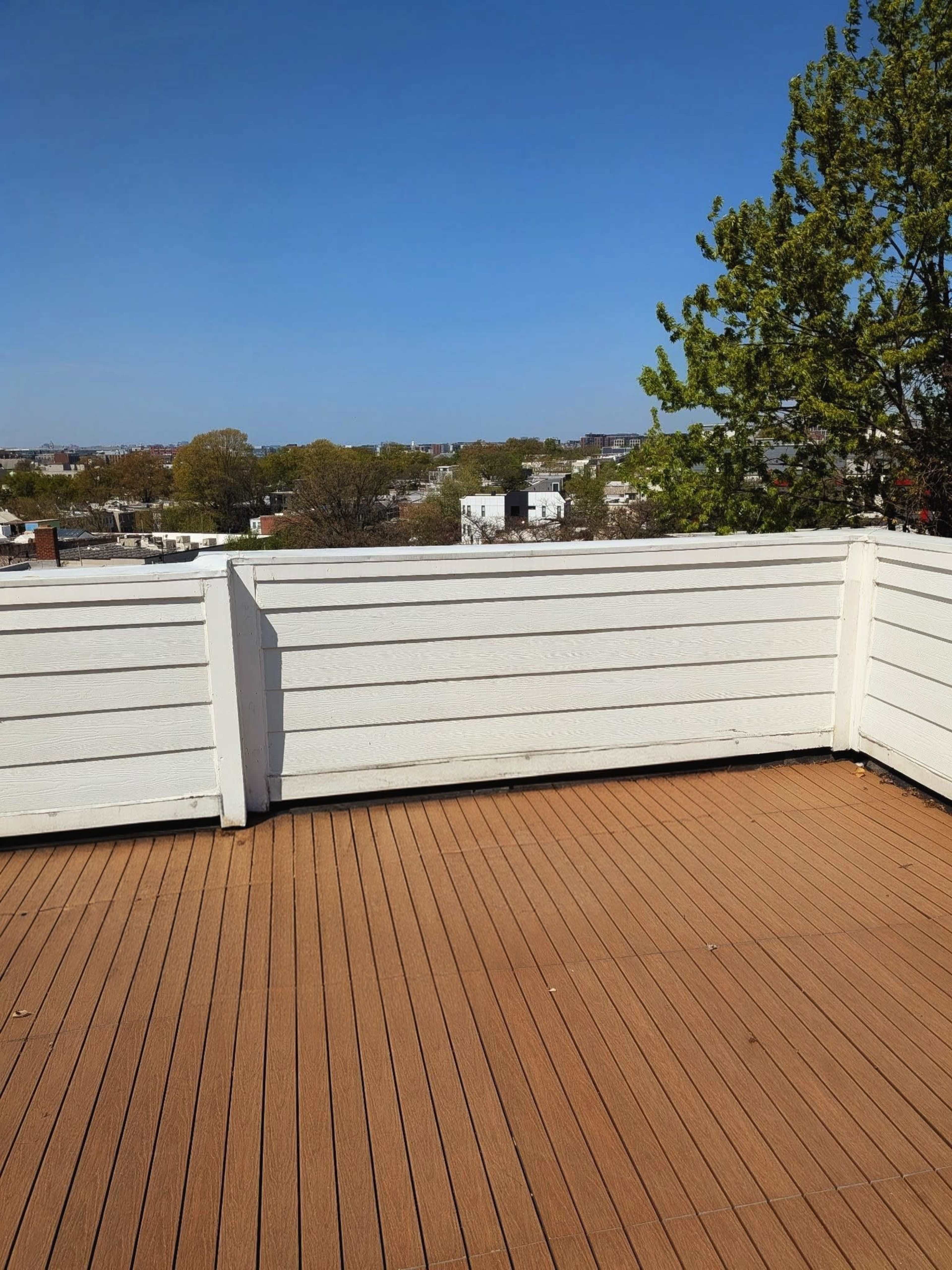 The image shows a wooden deck surrounded by a white railing, overlooking a view of buildings and trees under a clear blue sky.