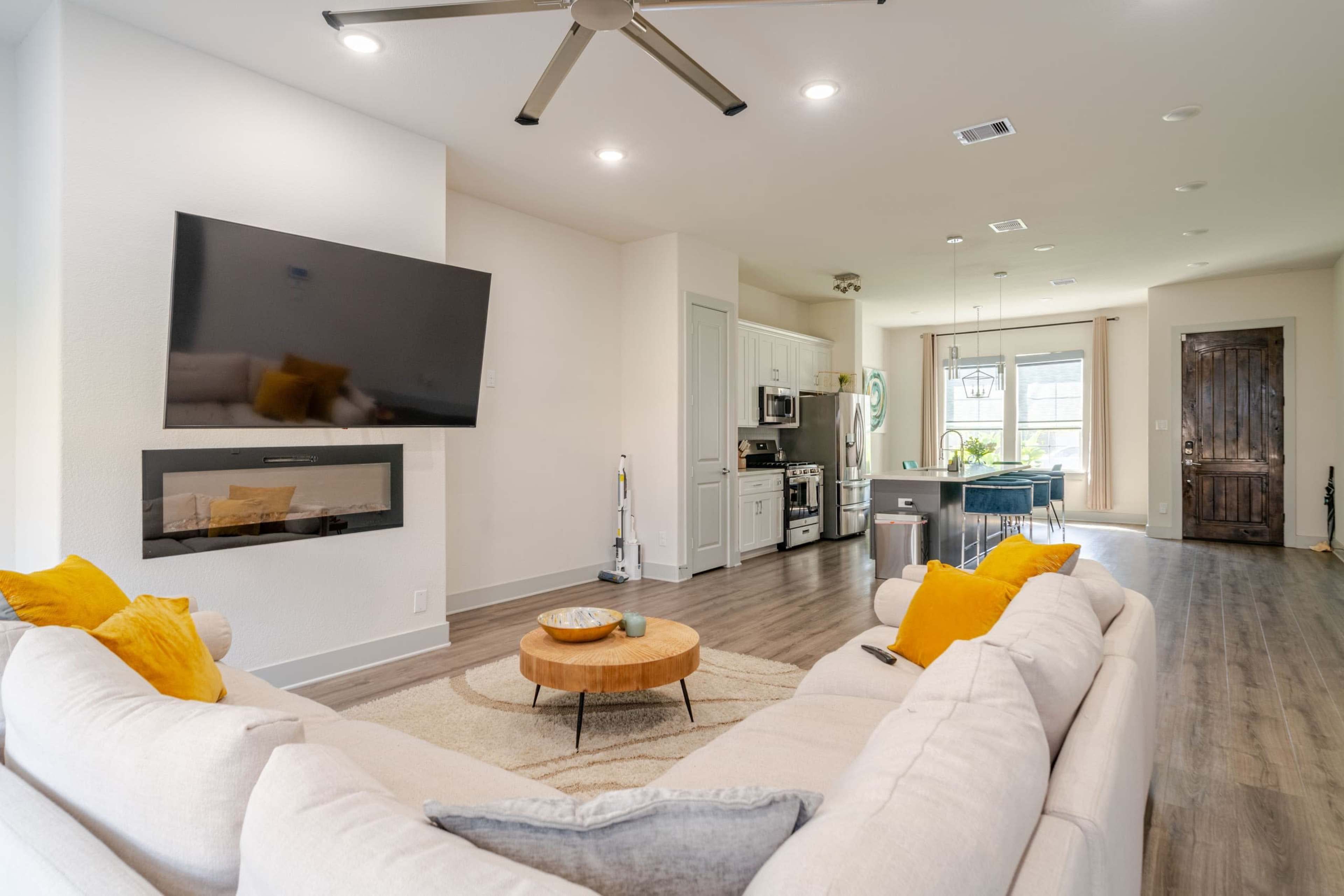The image shows a modern living room with a light-colored sectional sofa facing a mounted television and a built-in fireplace, adjacent to a kitchen area with a dining table and large windows for natural light.