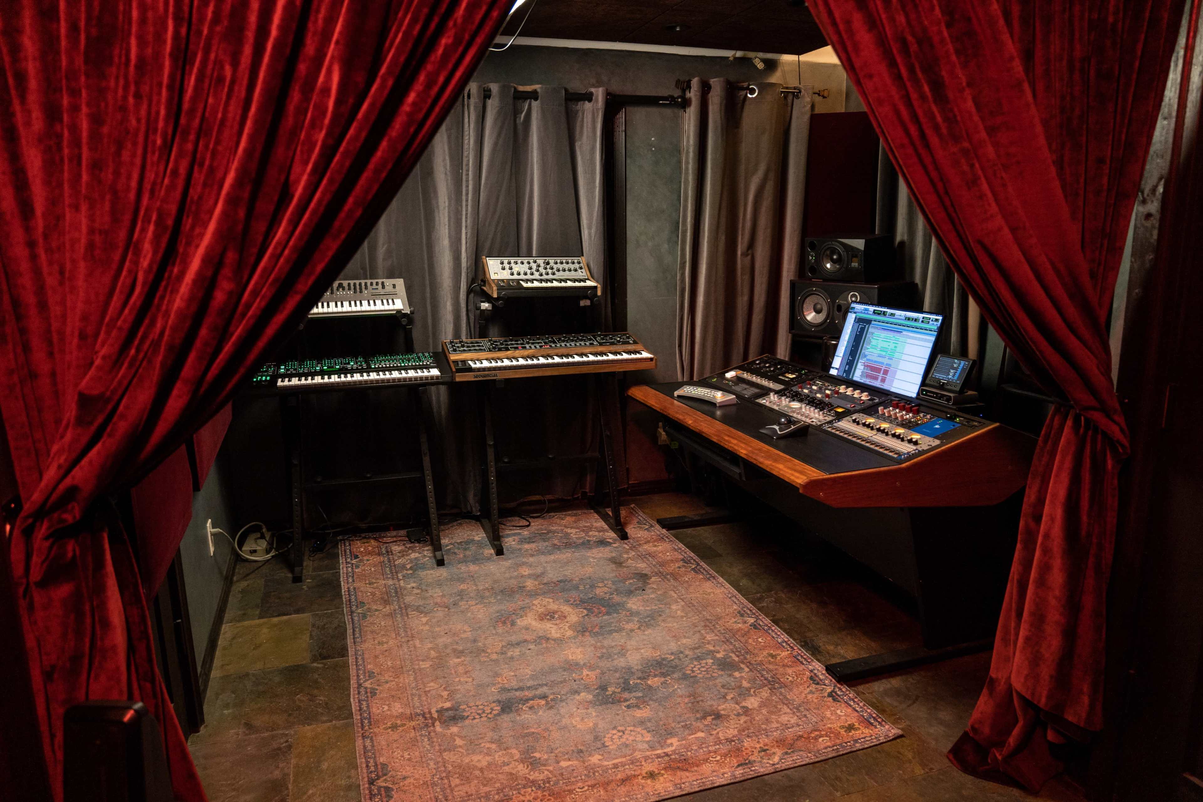 A music production studio featuring synthesizers on two stands, a mixing console with a screen, and red curtains framing the entrance.