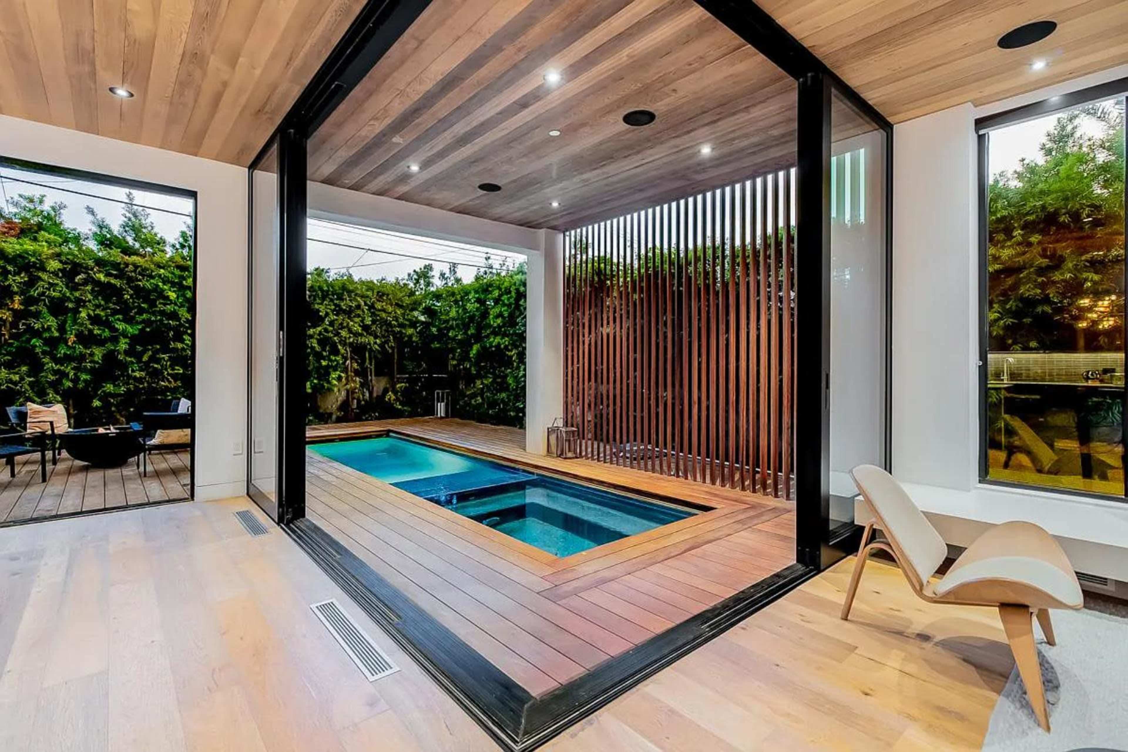 The image shows a modern indoor pool area with large sliding glass doors, wooden ceilings, and surrounding greenery.