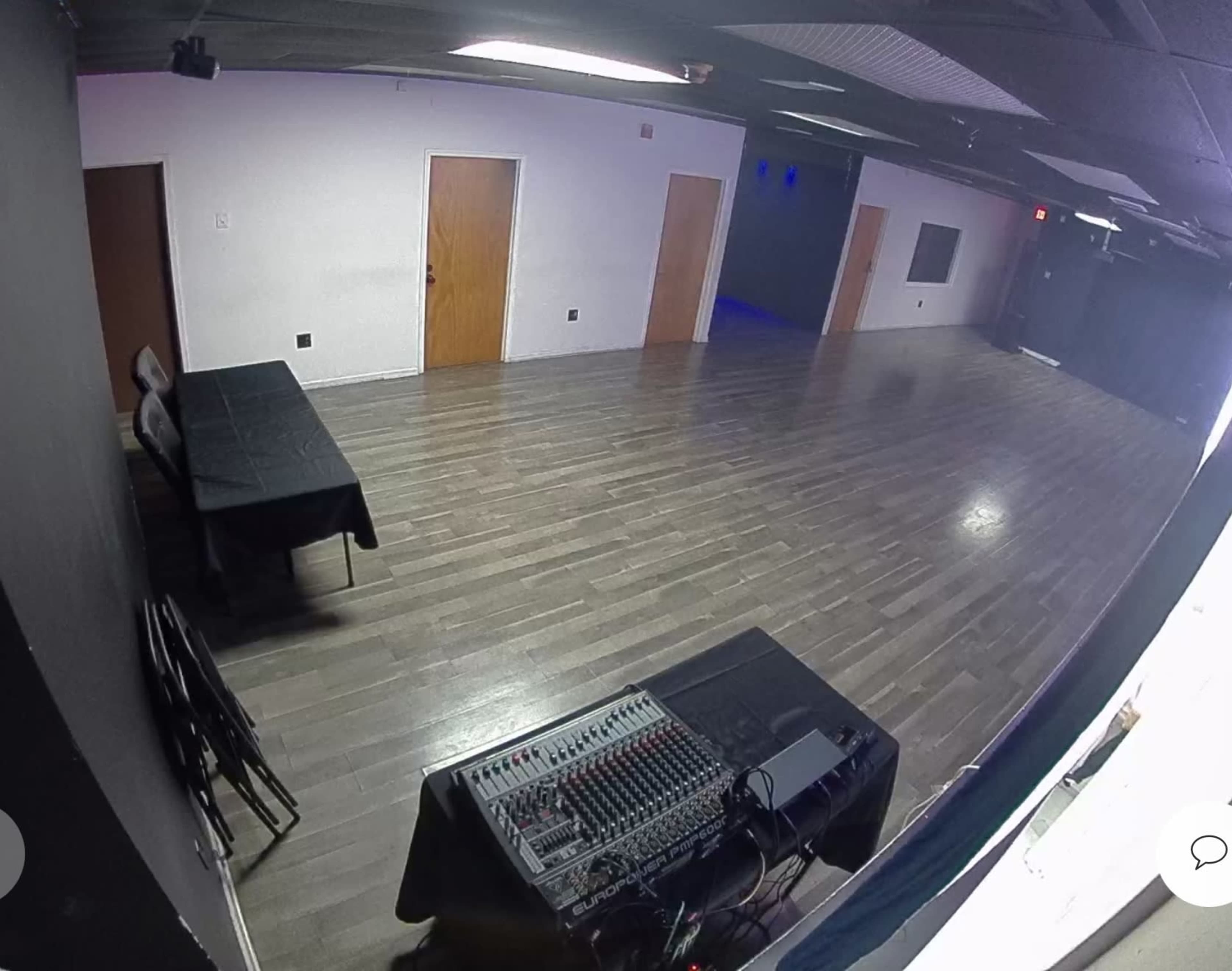 The image shows a large, empty room with polished wooden floors, a sound mixer on a table, and several closed doors along the walls.