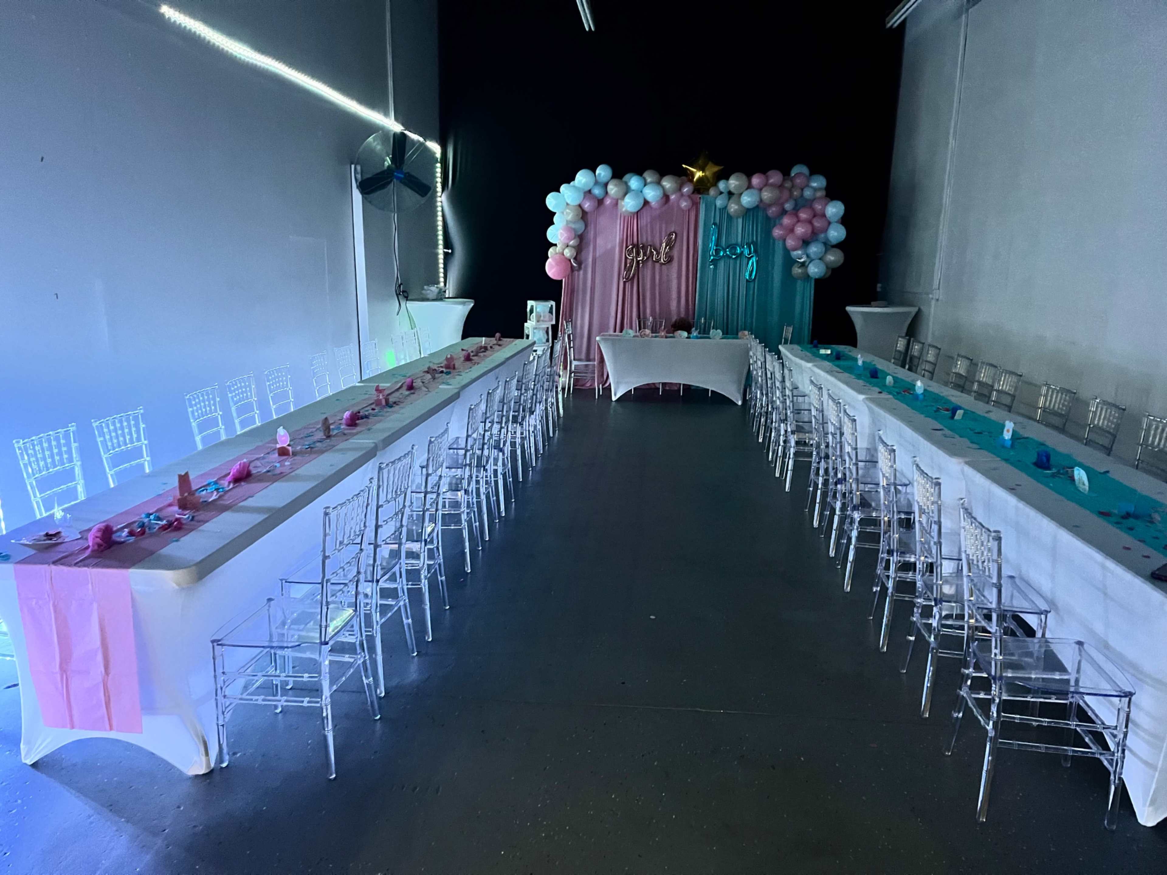 A spacious indoor venue features two long tables decorated with pink and blue accents, flanked by clear acrylic chairs, with a backdrop of balloons and a decorative stand at the front.