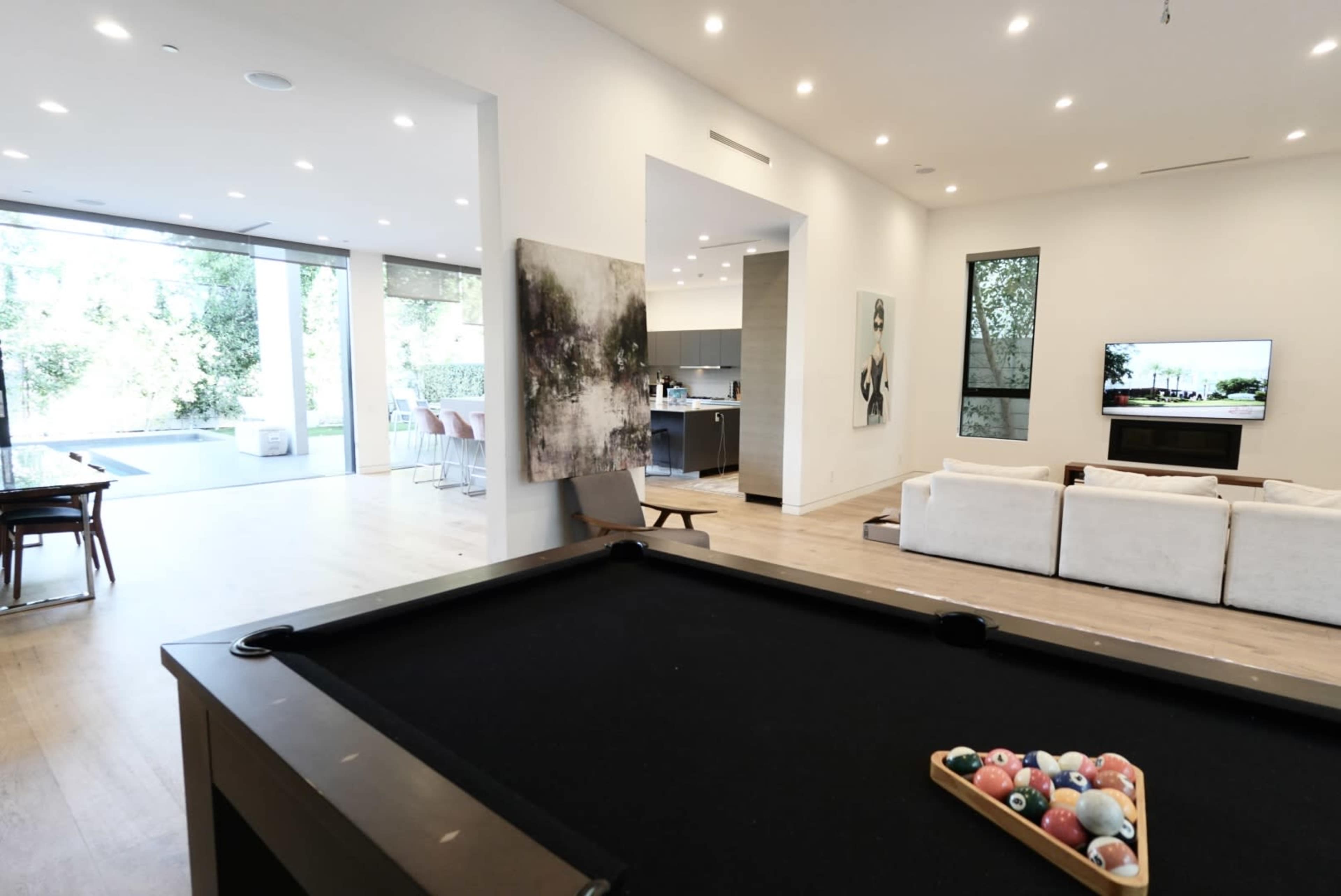 A pool table is in the foreground, overlooking a modern living space with large windows revealing an outdoor area.