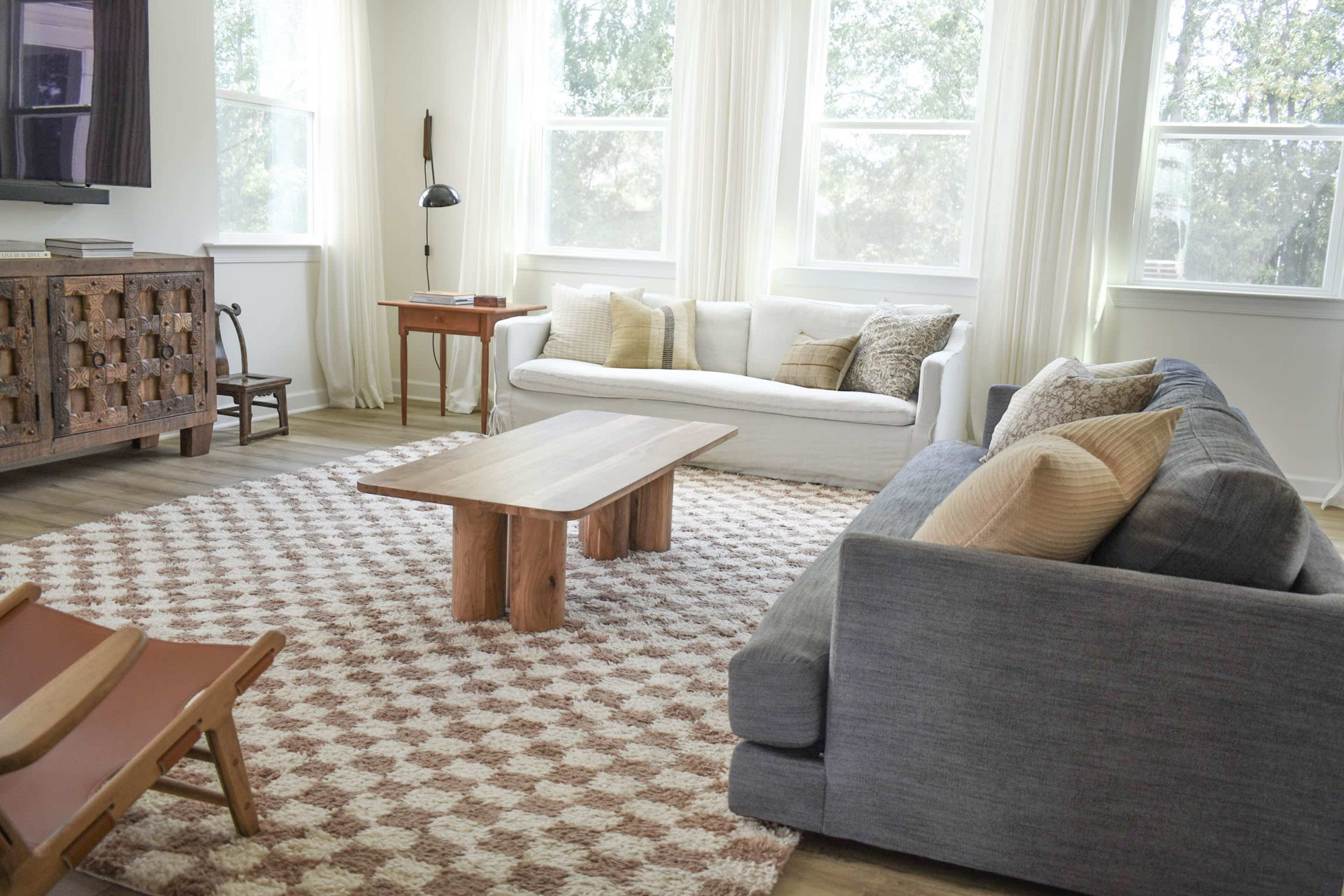 A cozy living room features a light-colored sofa, a gray sofa, a large wooden coffee table, and a patterned area rug, with ample natural light coming through large windows.