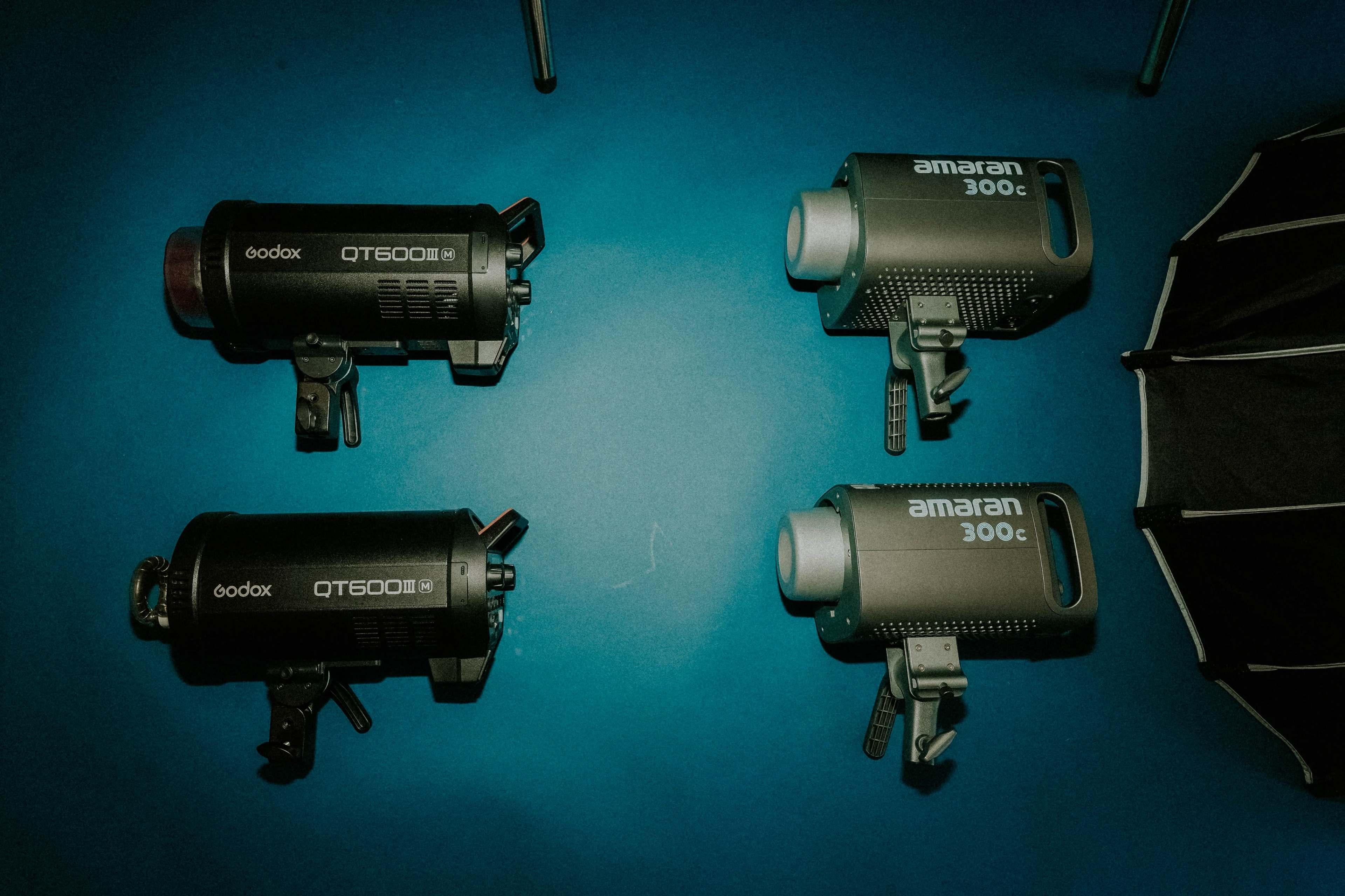 The image shows two Godox QT600II lights and two Amaran 300c lights arranged on a blue surface, accompanied by a softbox umbrella.