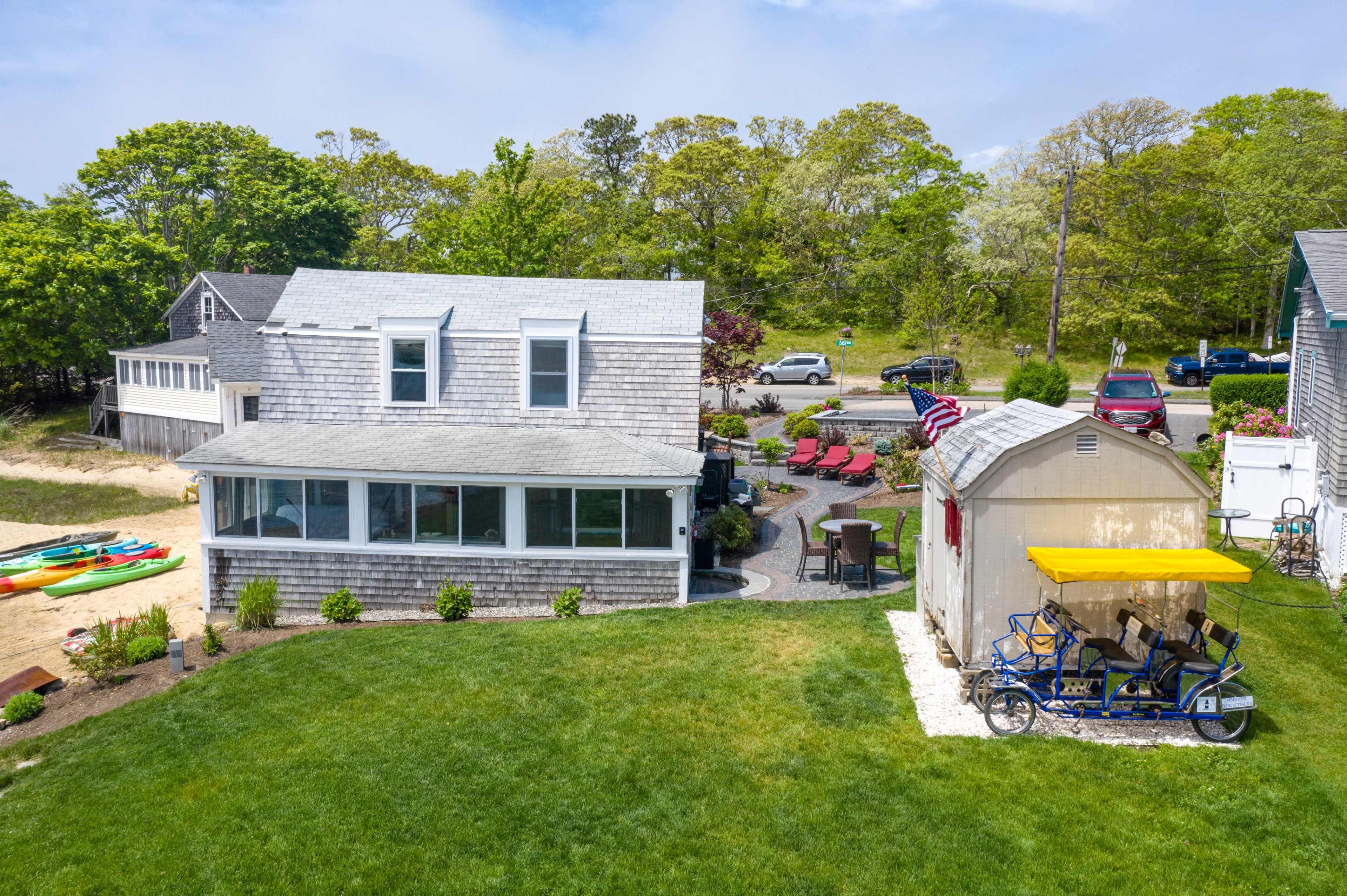 A gray-shingled house with large windows sits in a yard surrounded by green grass, patio chairs, and a storage shed with a yellow roof.
