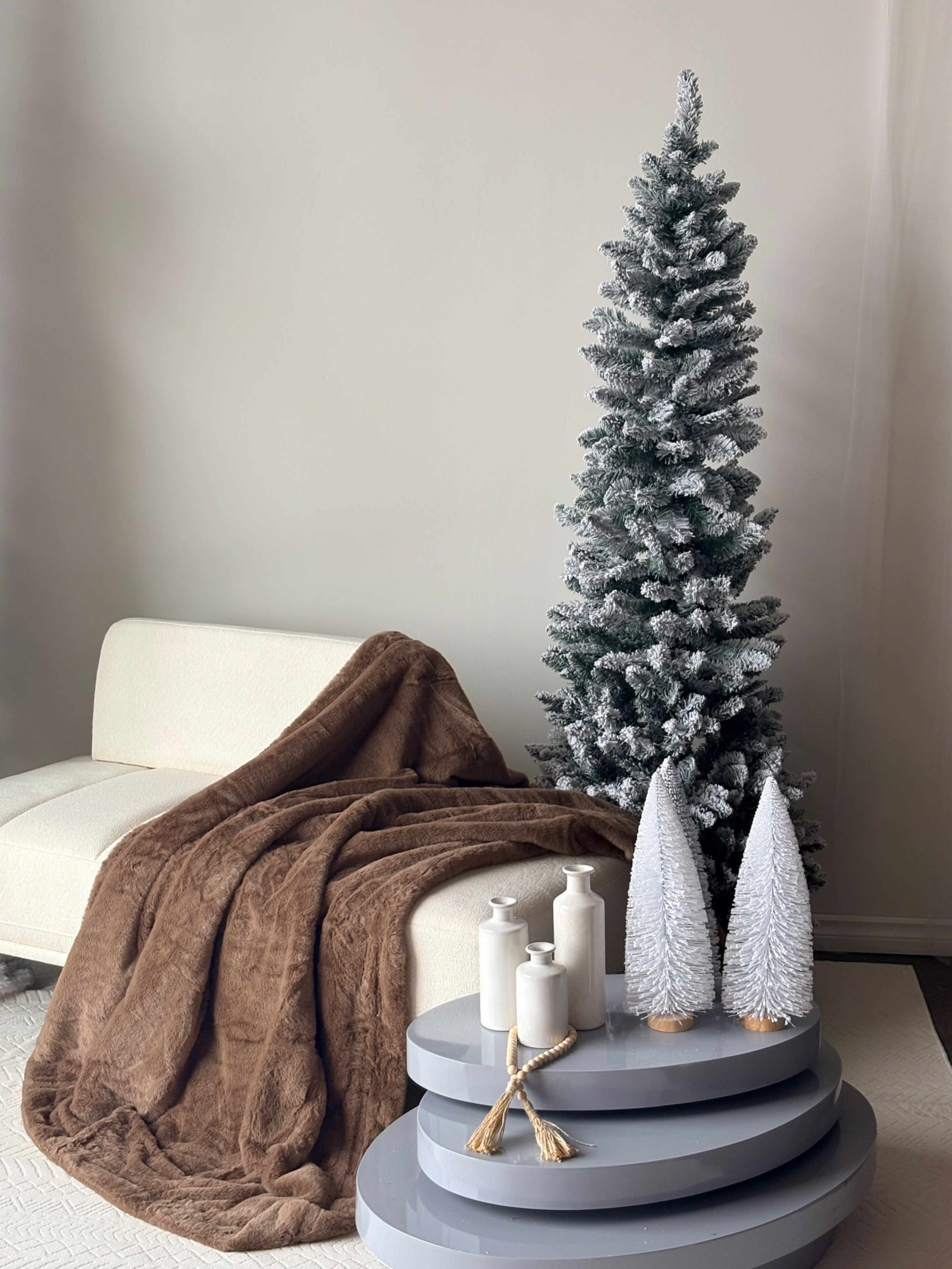 A small, frosted Christmas tree stands next to a white couch draped with a brown blanket, accompanied by candles and decorative trees on a gray pedestal.