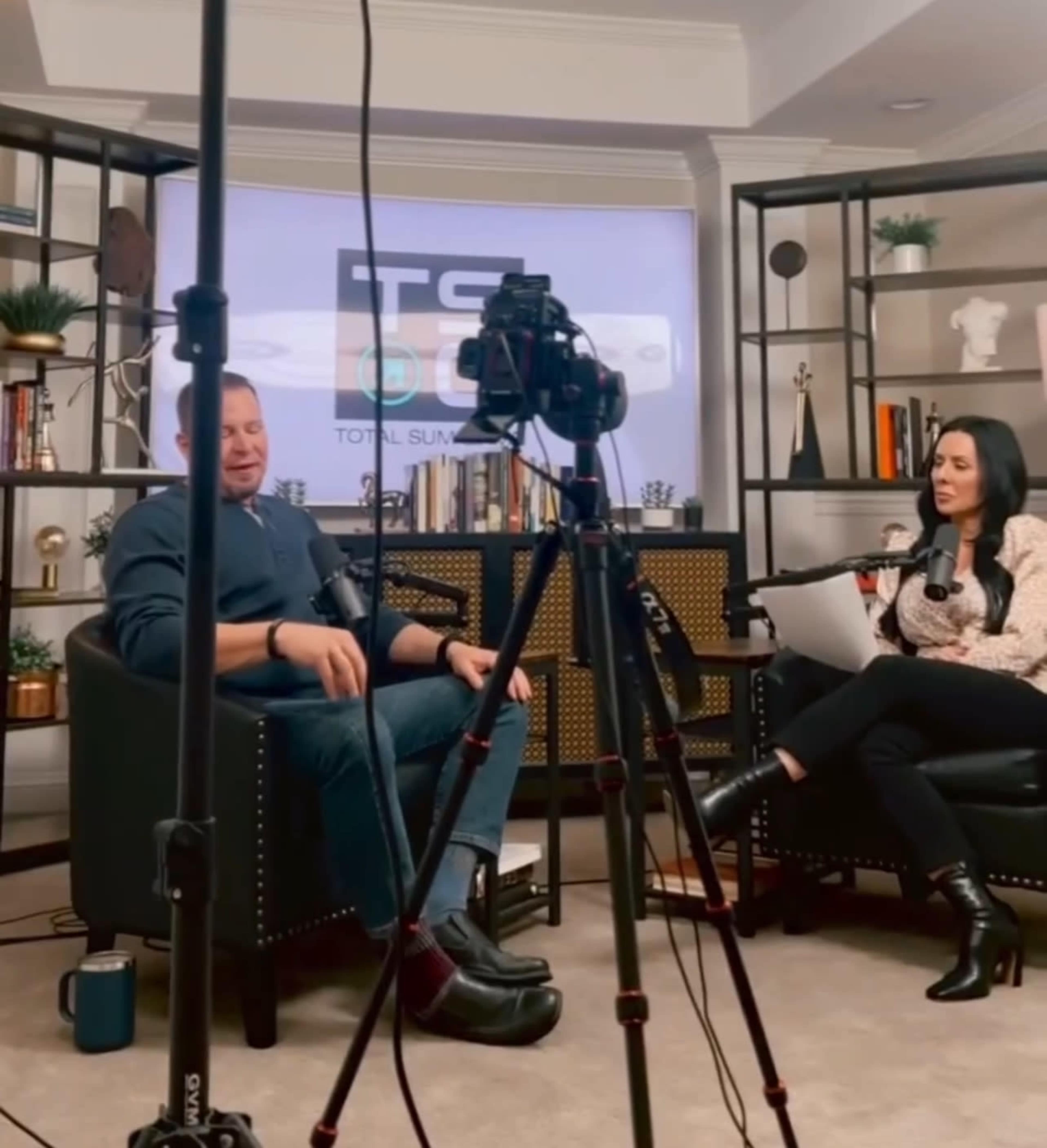 A man and a woman are seated on separate chairs in a well-lit room, engaging in a conversation while a camera is positioned in front of them.