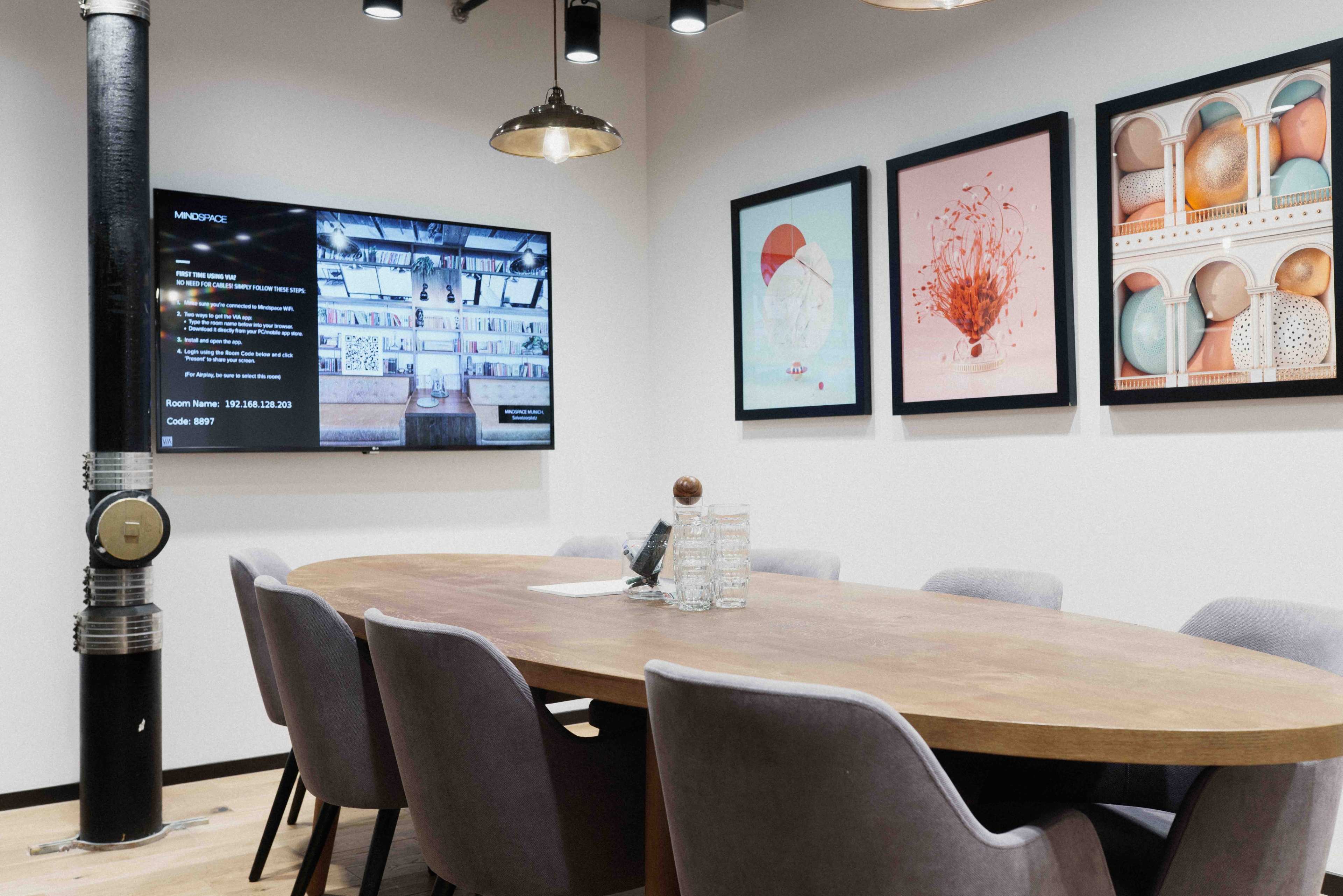 A meeting room with a large wooden table surrounded by upholstered chairs, a television displaying information on the wall, and framed artwork hanging nearby.