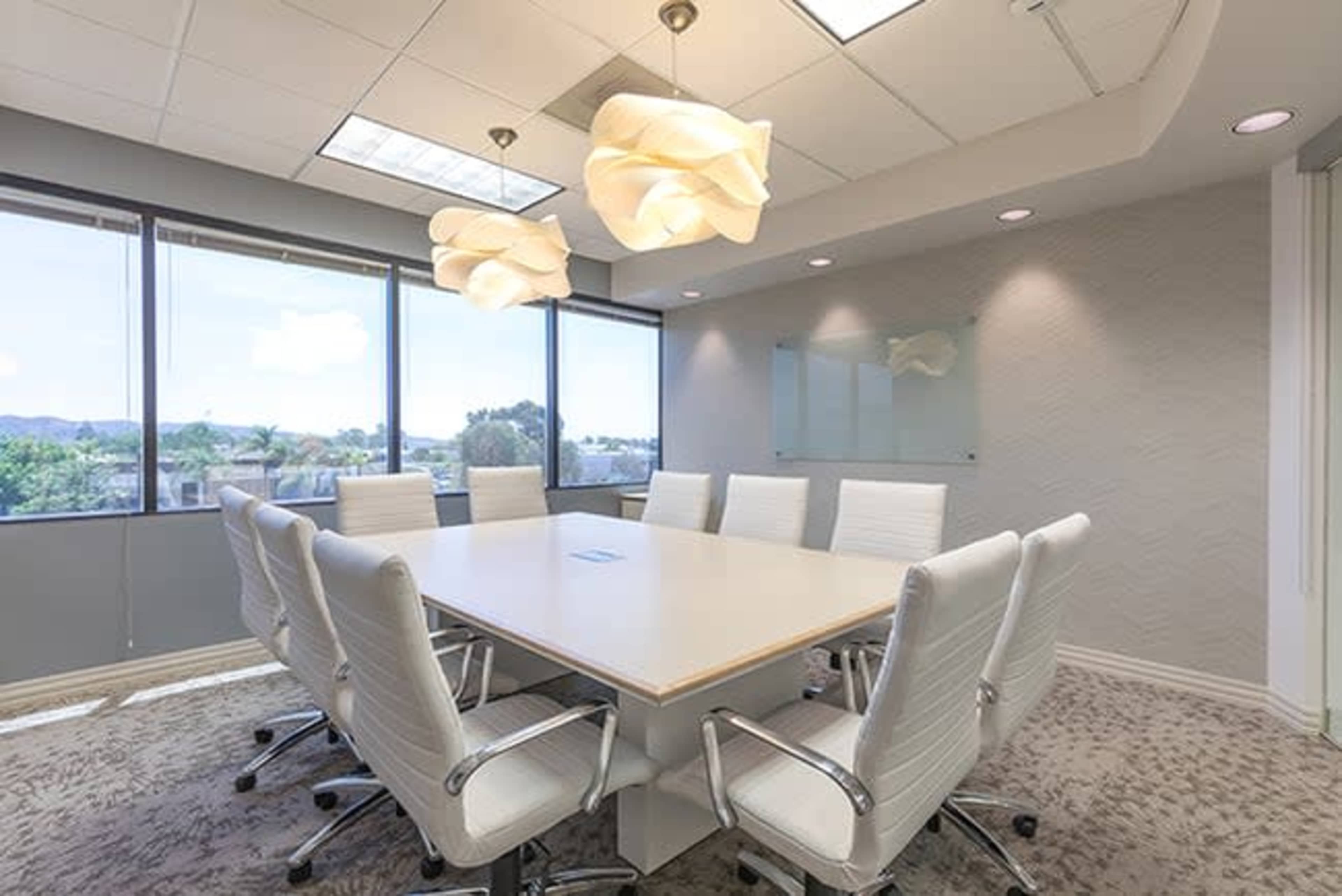 A modern conference room features a large table surrounded by white chairs, with large windows allowing natural light to enter.