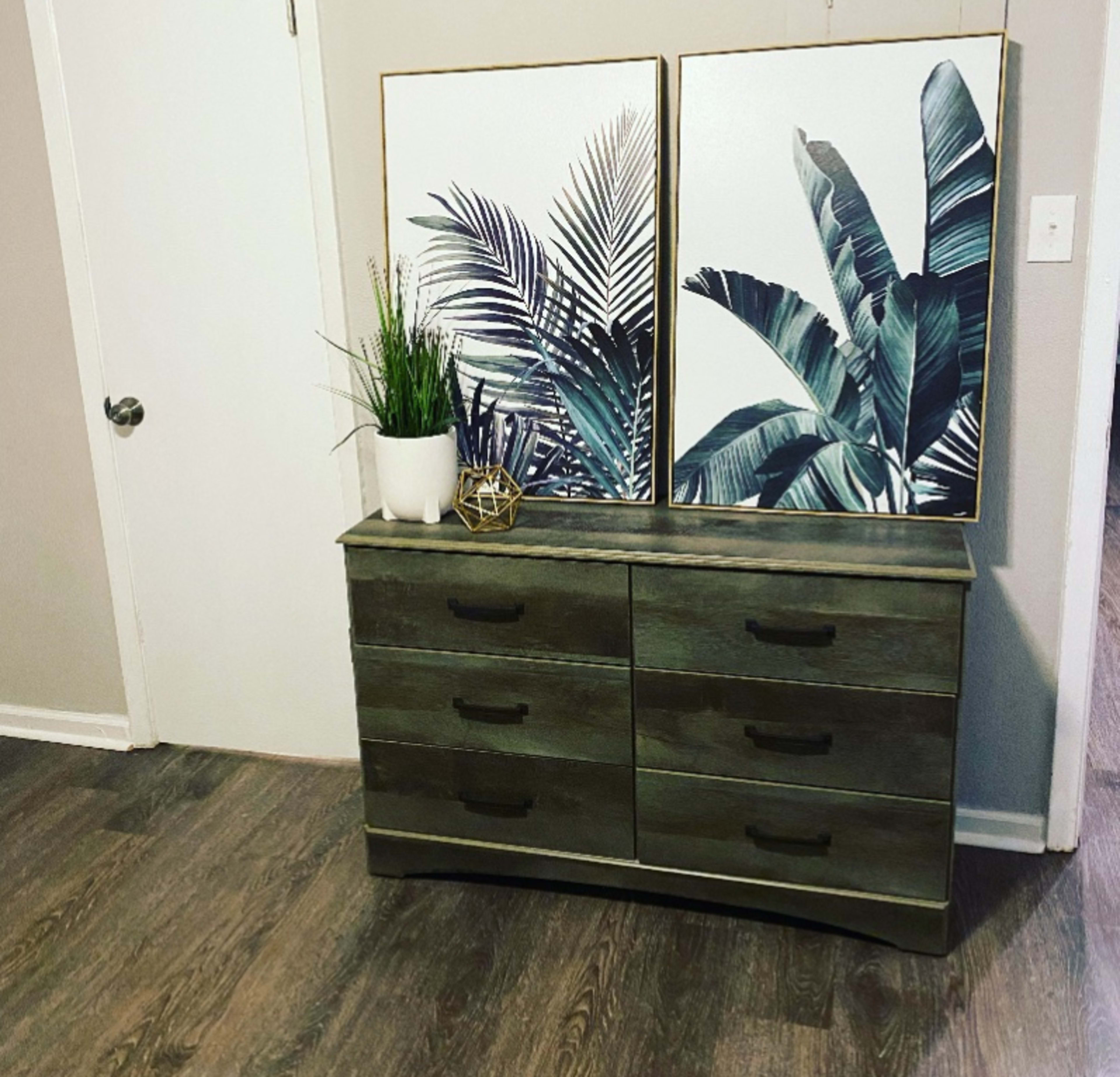 The image shows a dresser with six drawers, topped by two framed botanical prints and a potted plant.