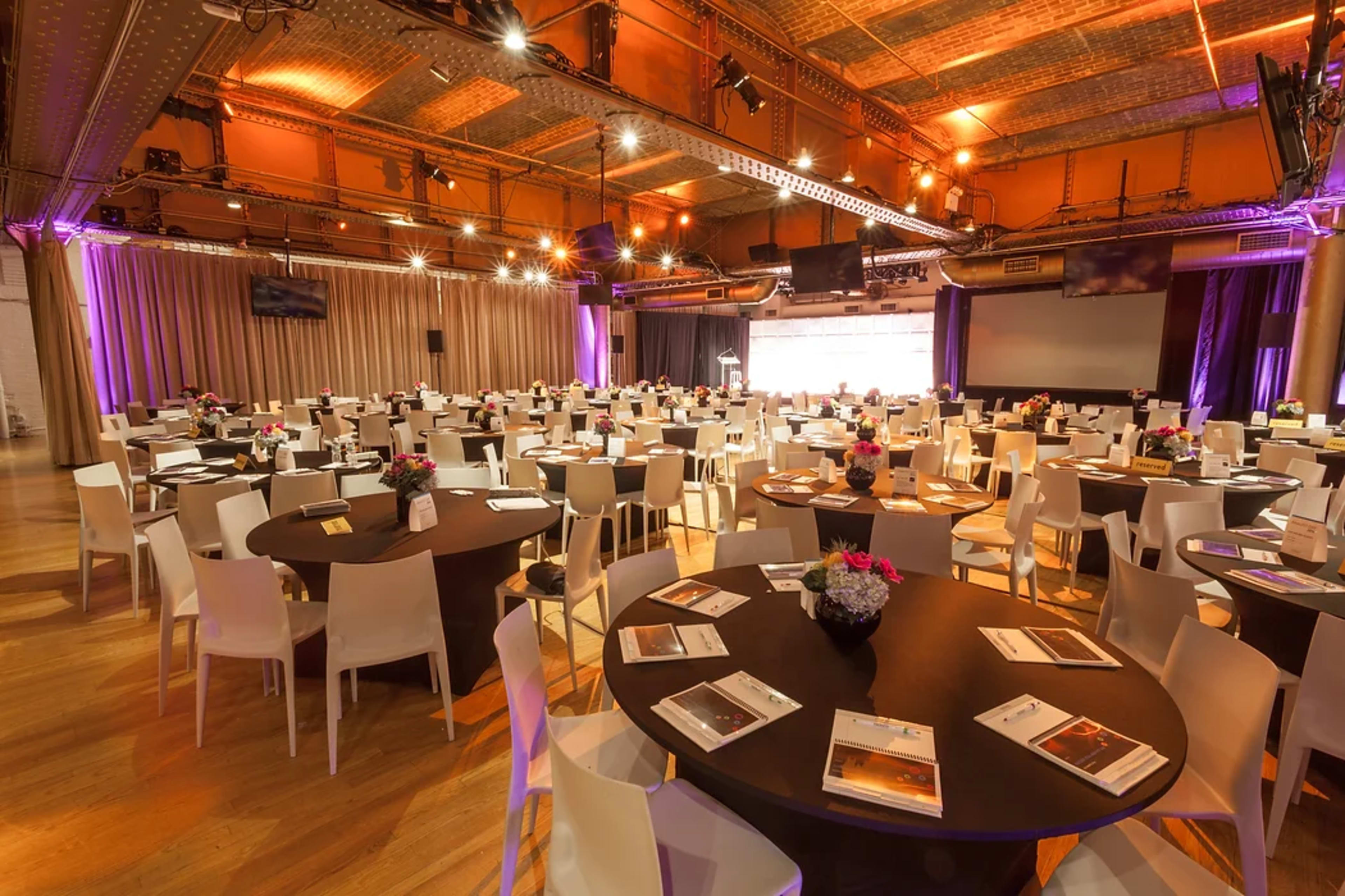 A spacious event venue is set up with round tables and chairs, arranged neatly for a formal gathering or dinner, under warm lighting.