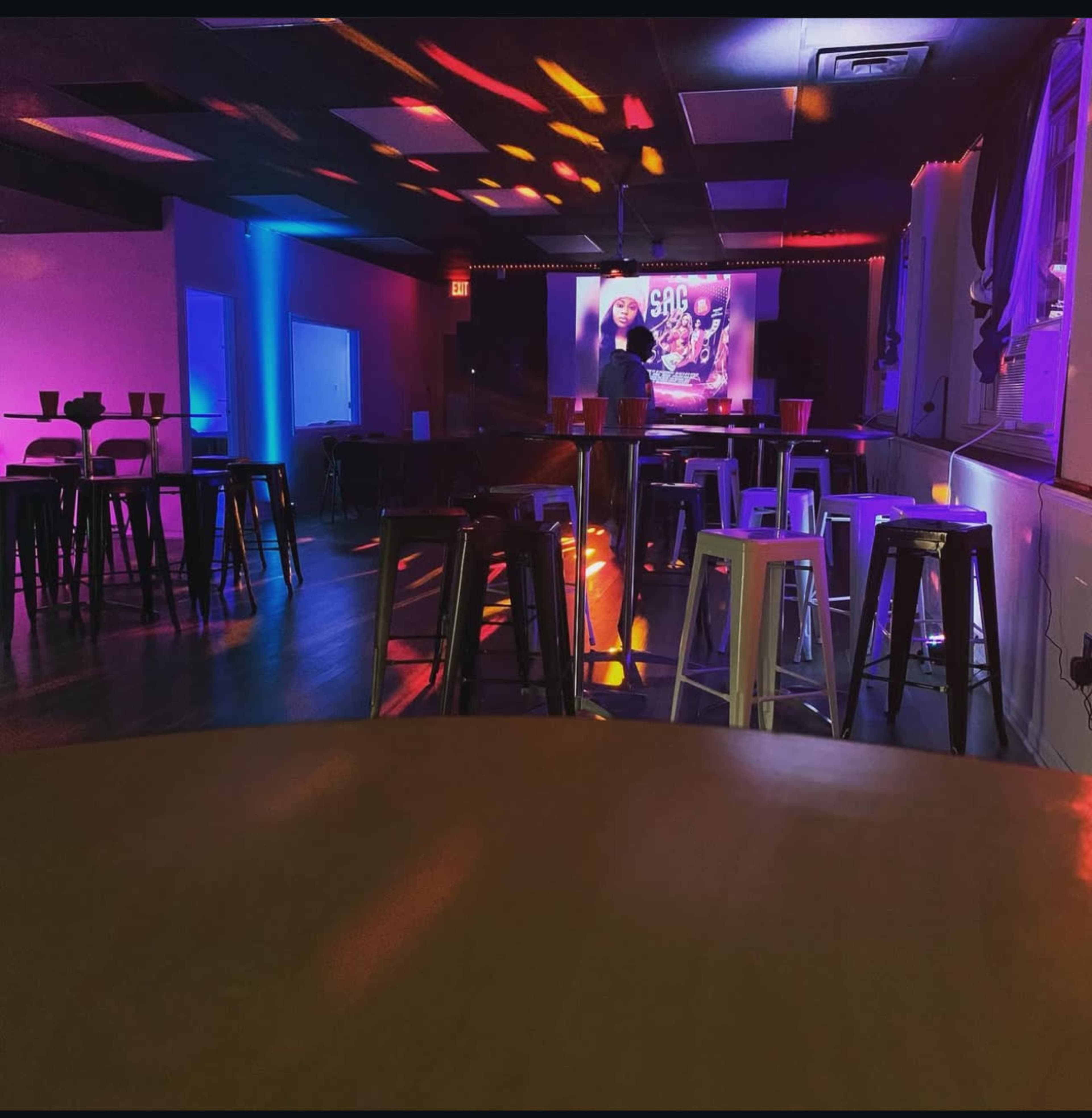 The image shows an empty bar or event space with colorful lighting and a large projector screen displaying a graphic in the background.